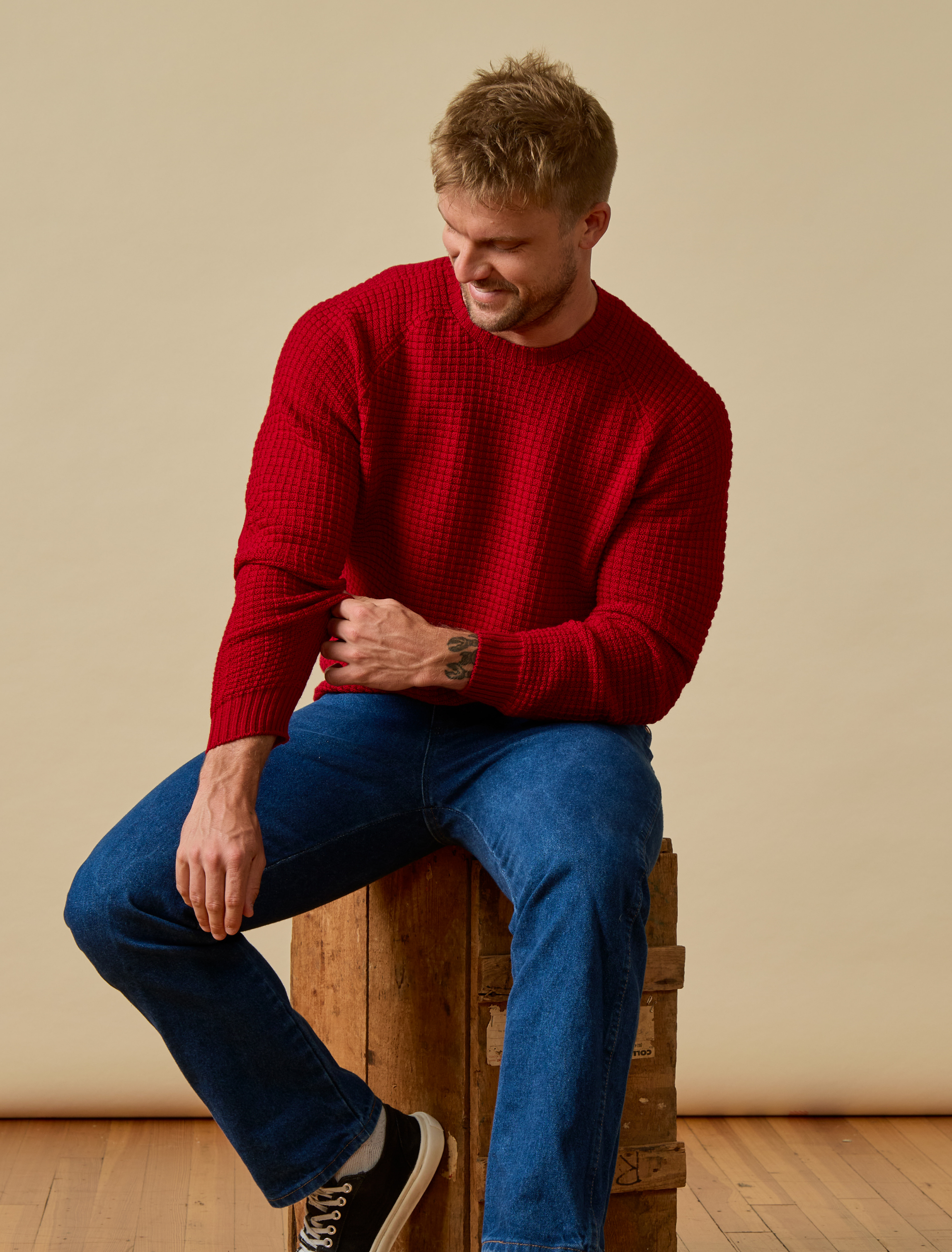 Man wearing a red Merino Wool sweater and blue jeans sitting on a wooden barrel against a beige wall.