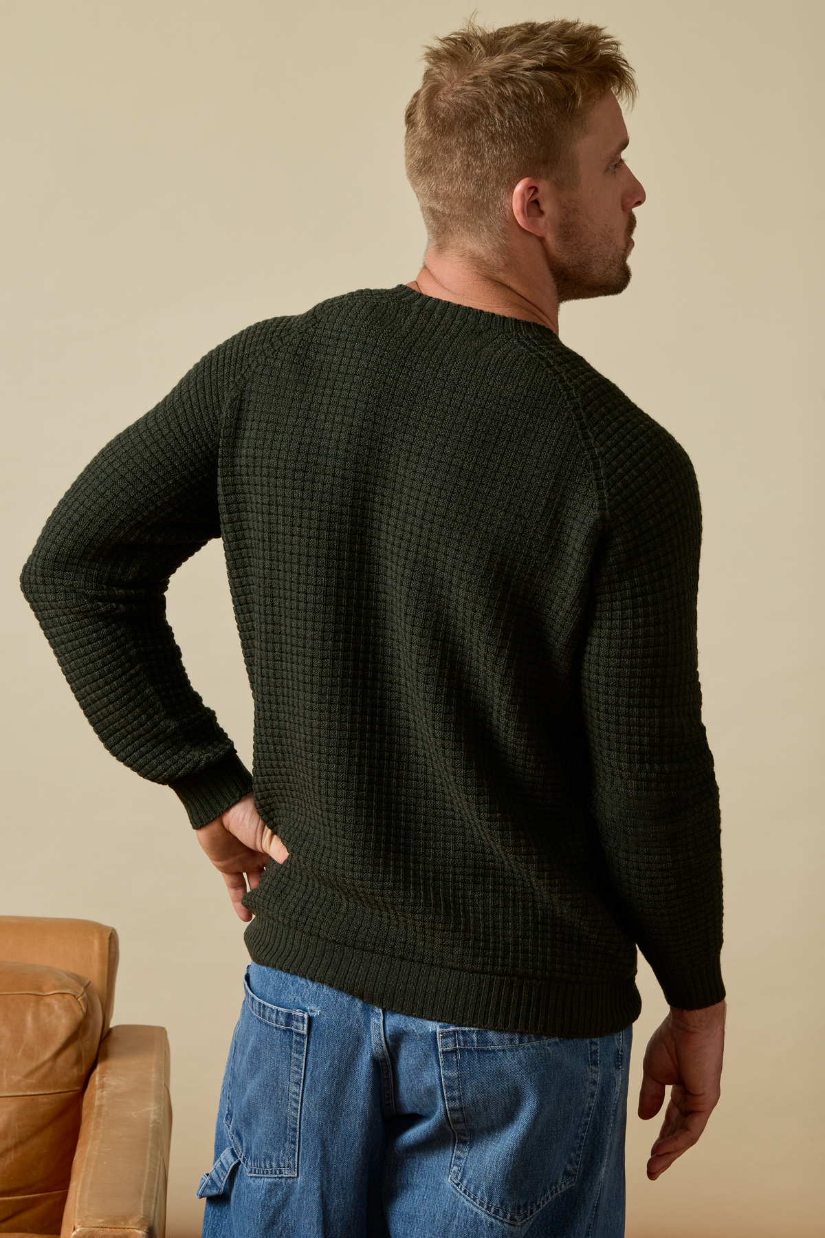 Man wearing a dark green Merino wool sweater and blue jeans standing against a beige wall. #color_forest green