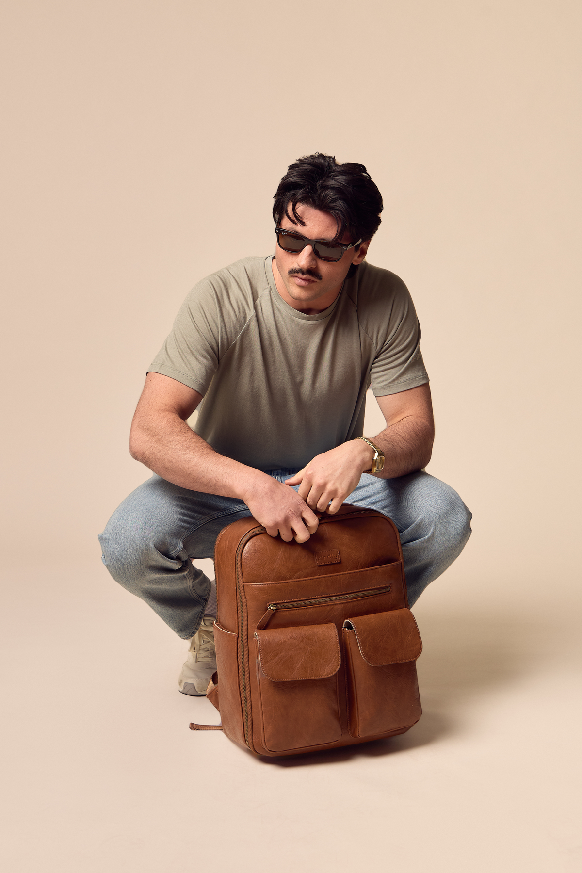 A man wearing sunglasses, a Cassius Merino Athletic t-shirt, and blue jeans crouches down next to a brown leather backpack on a solid beige background.
