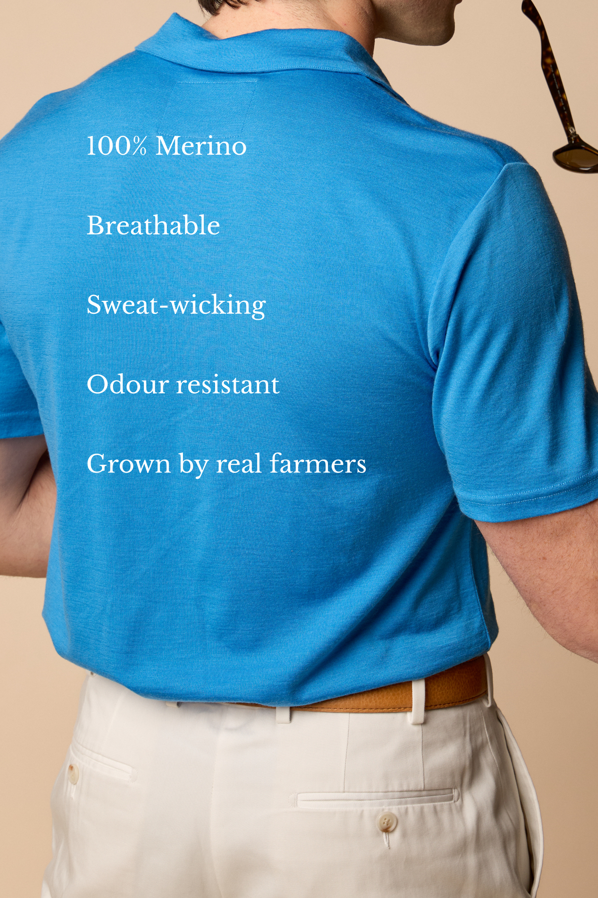 Person wearing a Sir W. Merino Bruce French Blue shirt with a logo, holding sunglasses, on a beige background #Color_French Blue