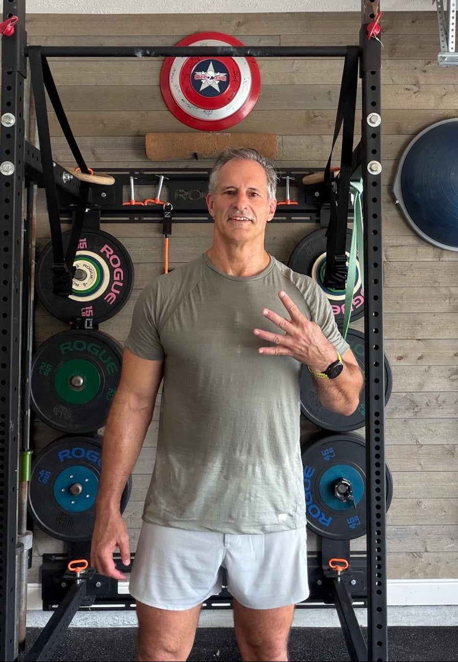 A man wearing a gray t-shirt and white shorts stands in front of a black power rack in a home gym setting, gesturing with his hand. The rack is loaded with weight plates and has a Captain America shield hanging on the wall behind it.