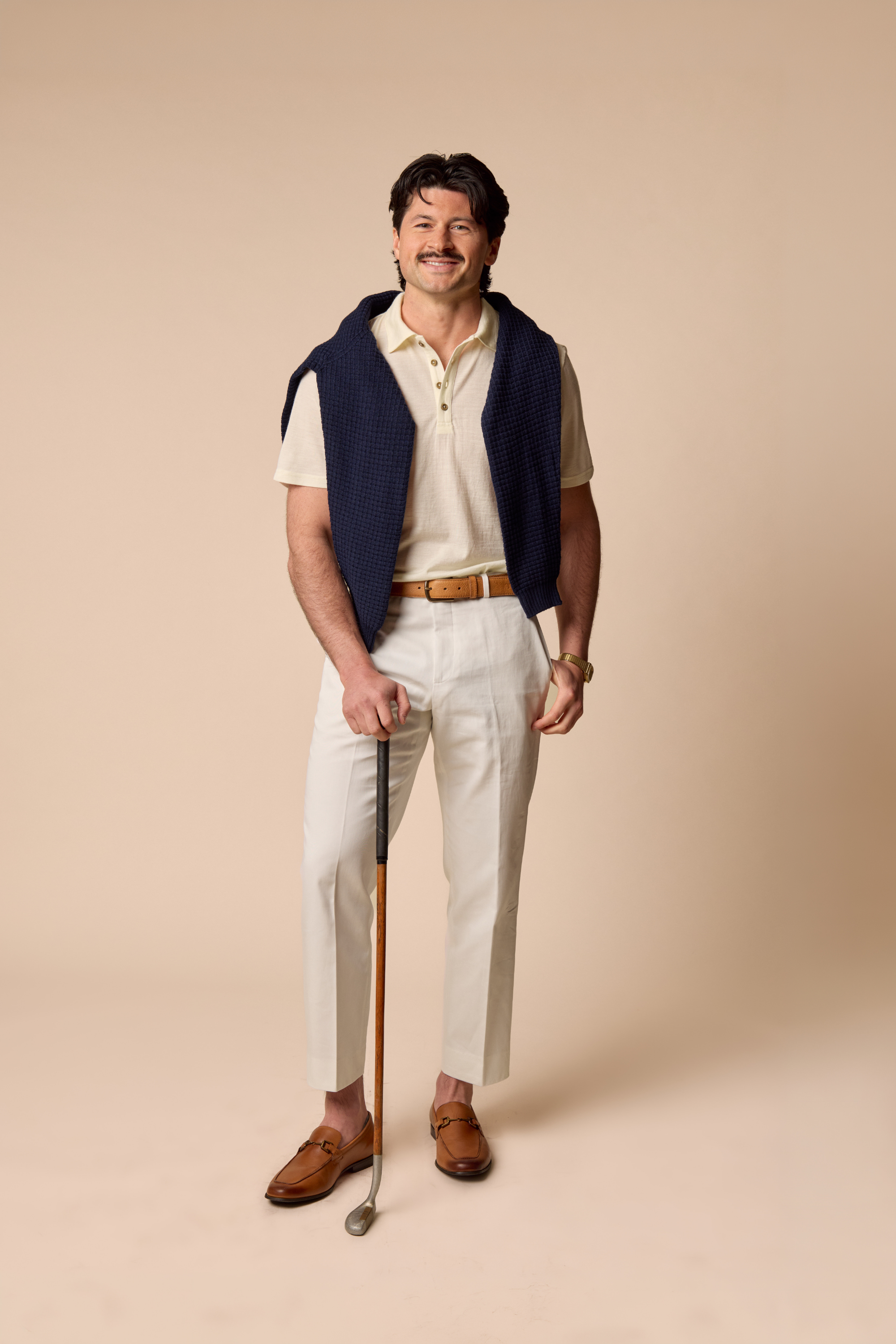 Man in a Sir W Natural Polo shirt with a navy vest and beige pants holding a golf club on a beige background