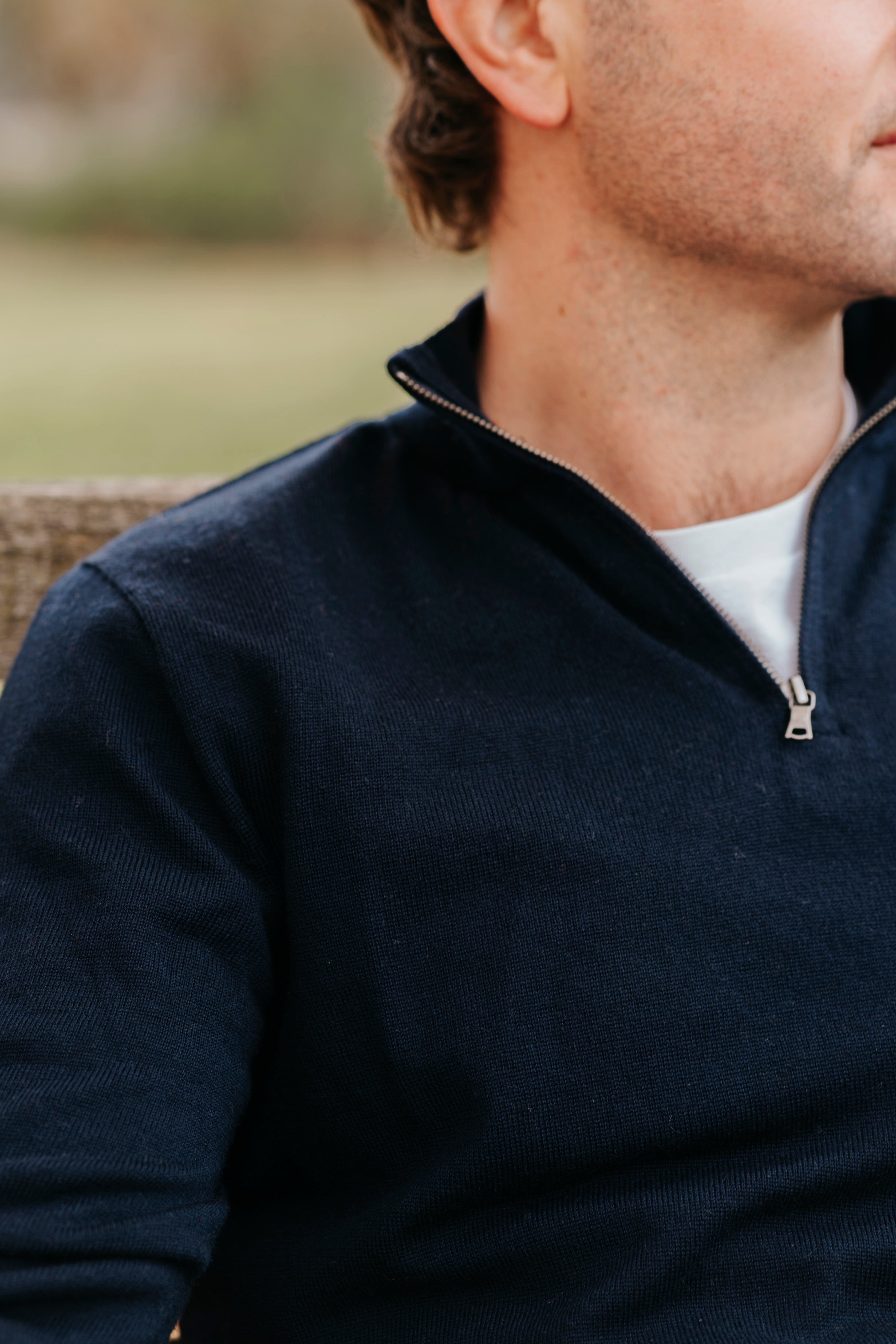 Man wearing a navy blue sweater with a white shirt underneath, sitting outdoors.