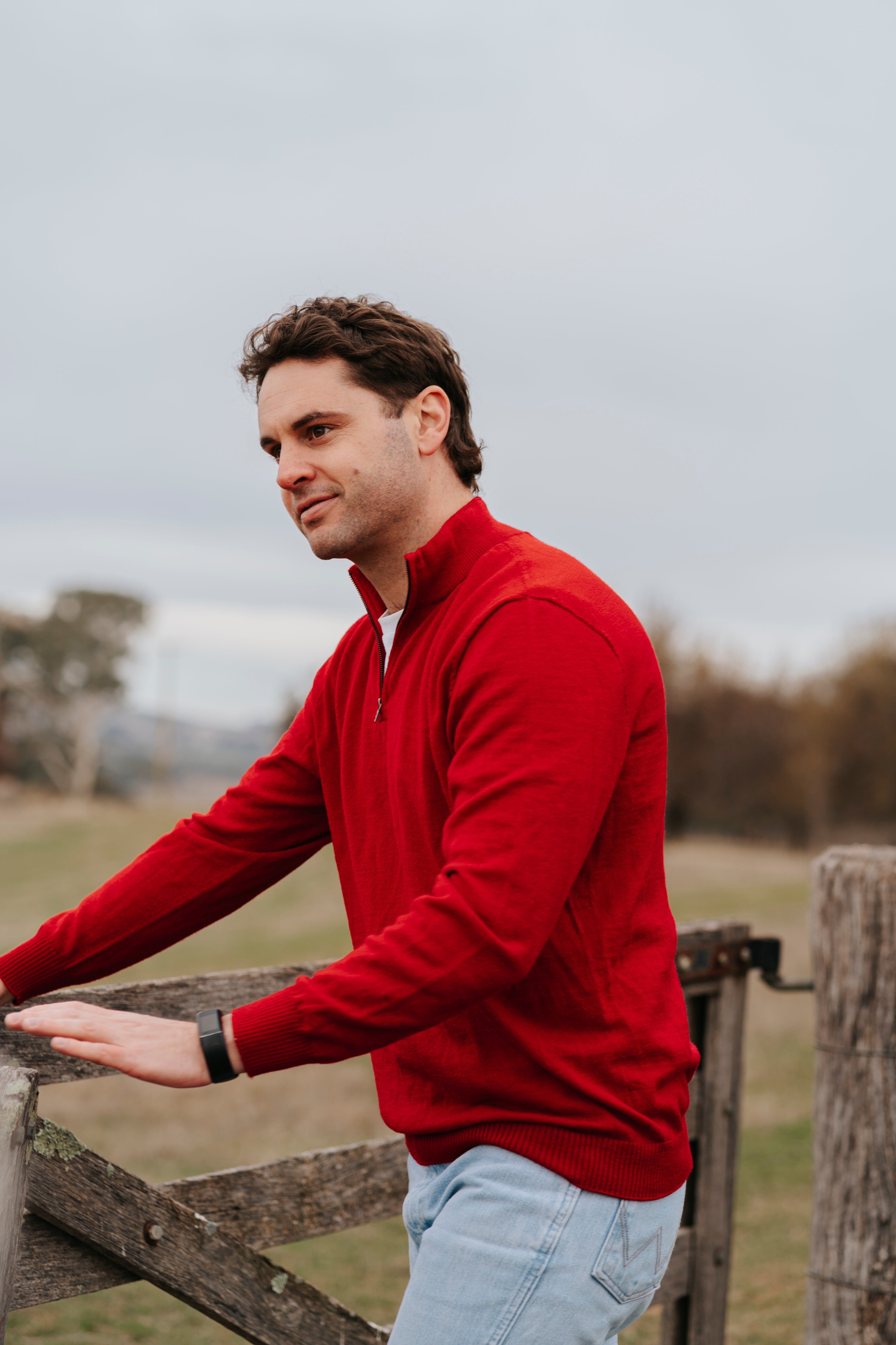 Man wearing a red merino sweater standing by a wooden fence in an outdoor setting
