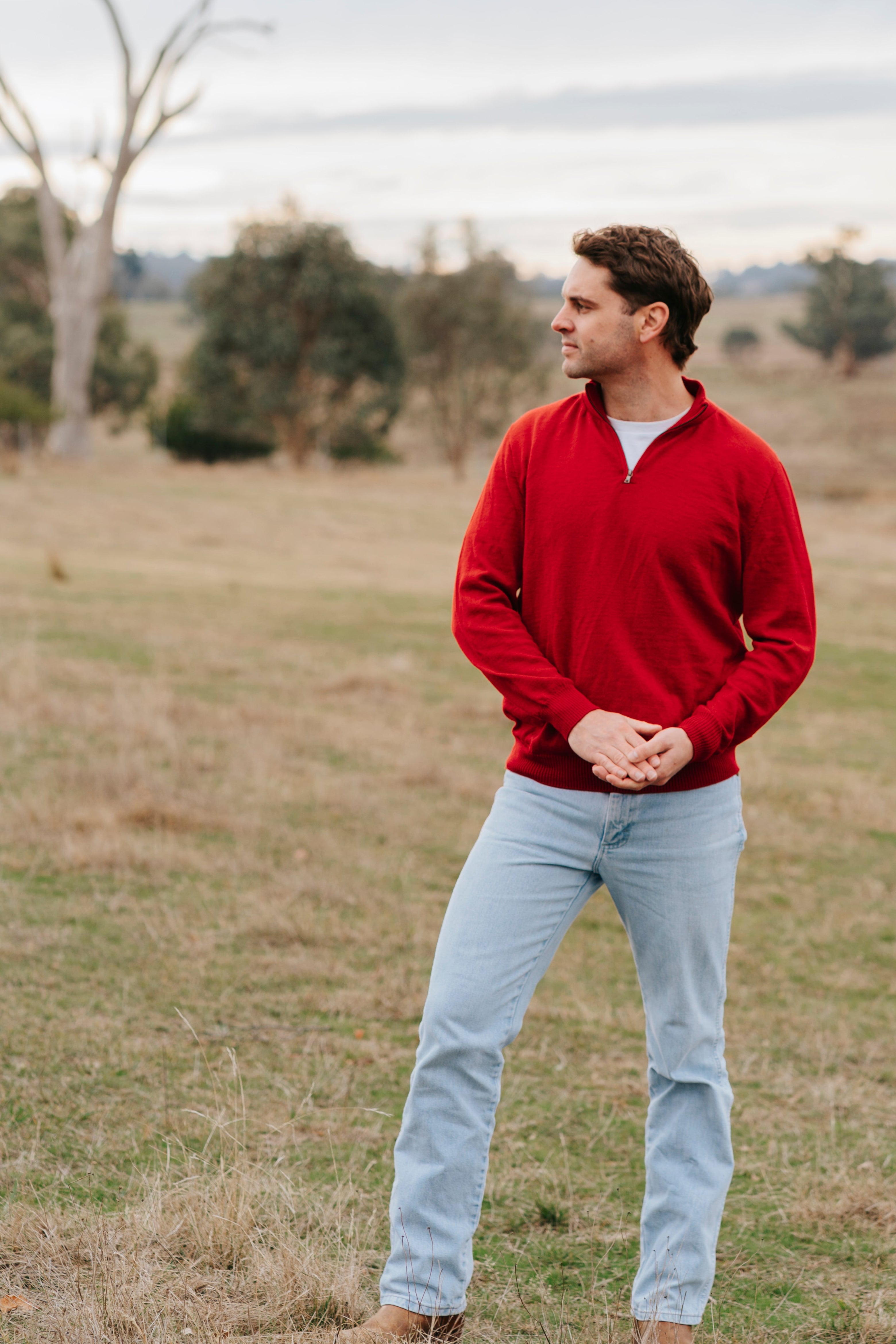 Man wearing a red sweater and light blue jeans standing in a field with trees in the background