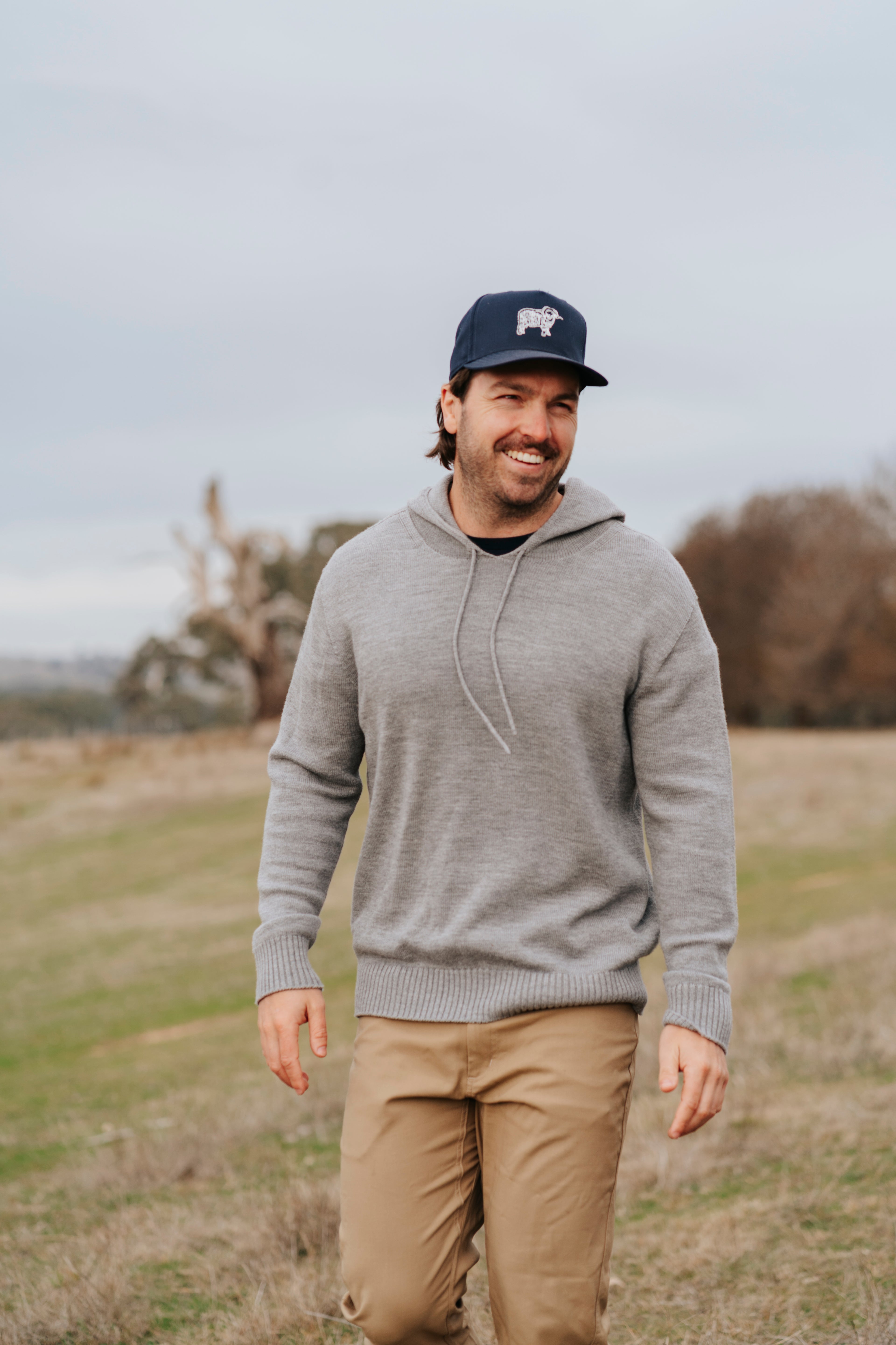 Man wearing a gray merino hoodie and navy Sir W. cap standing in a field