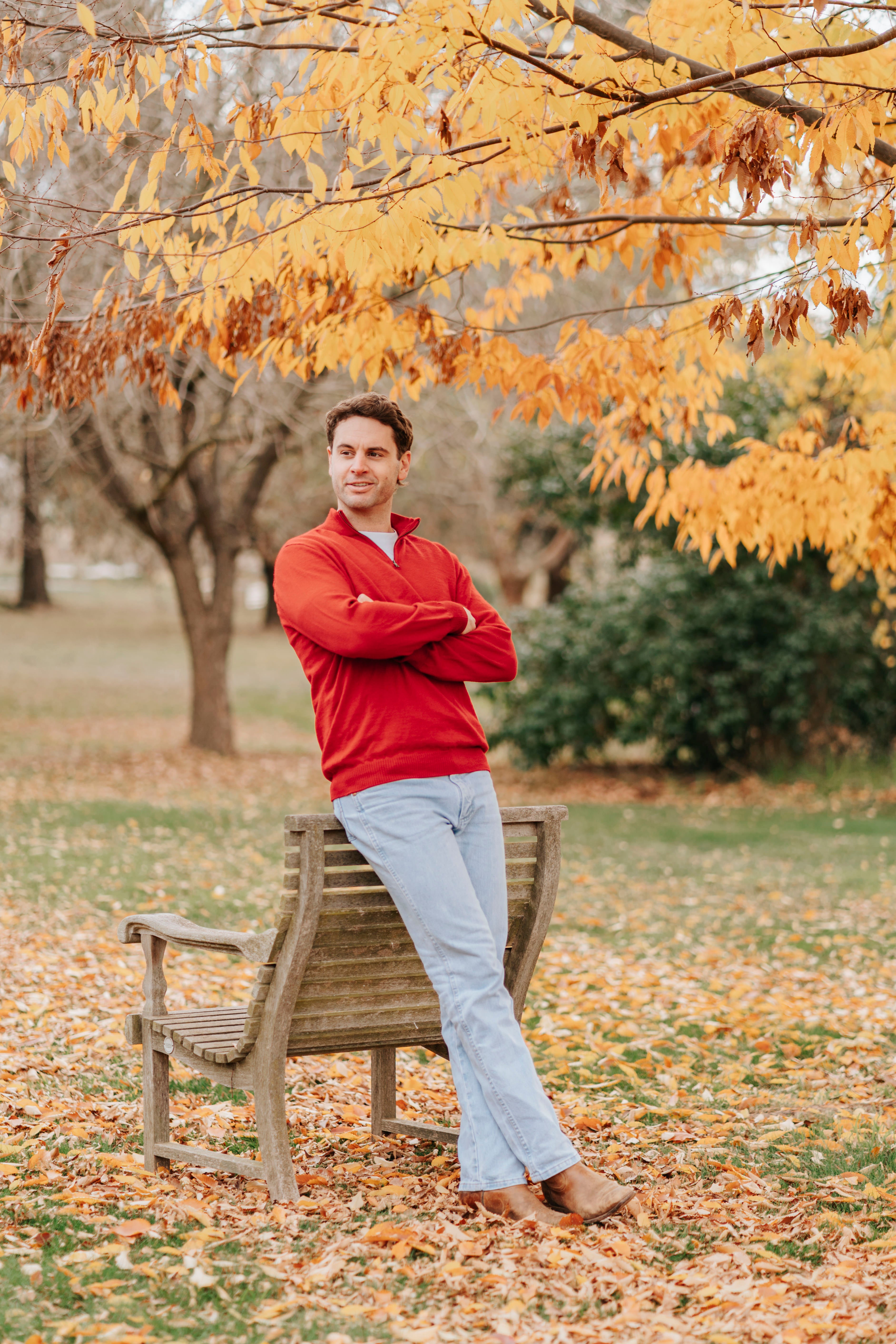 Man in a red merino sweater standing next to a bench under a tree with autumn leaves.