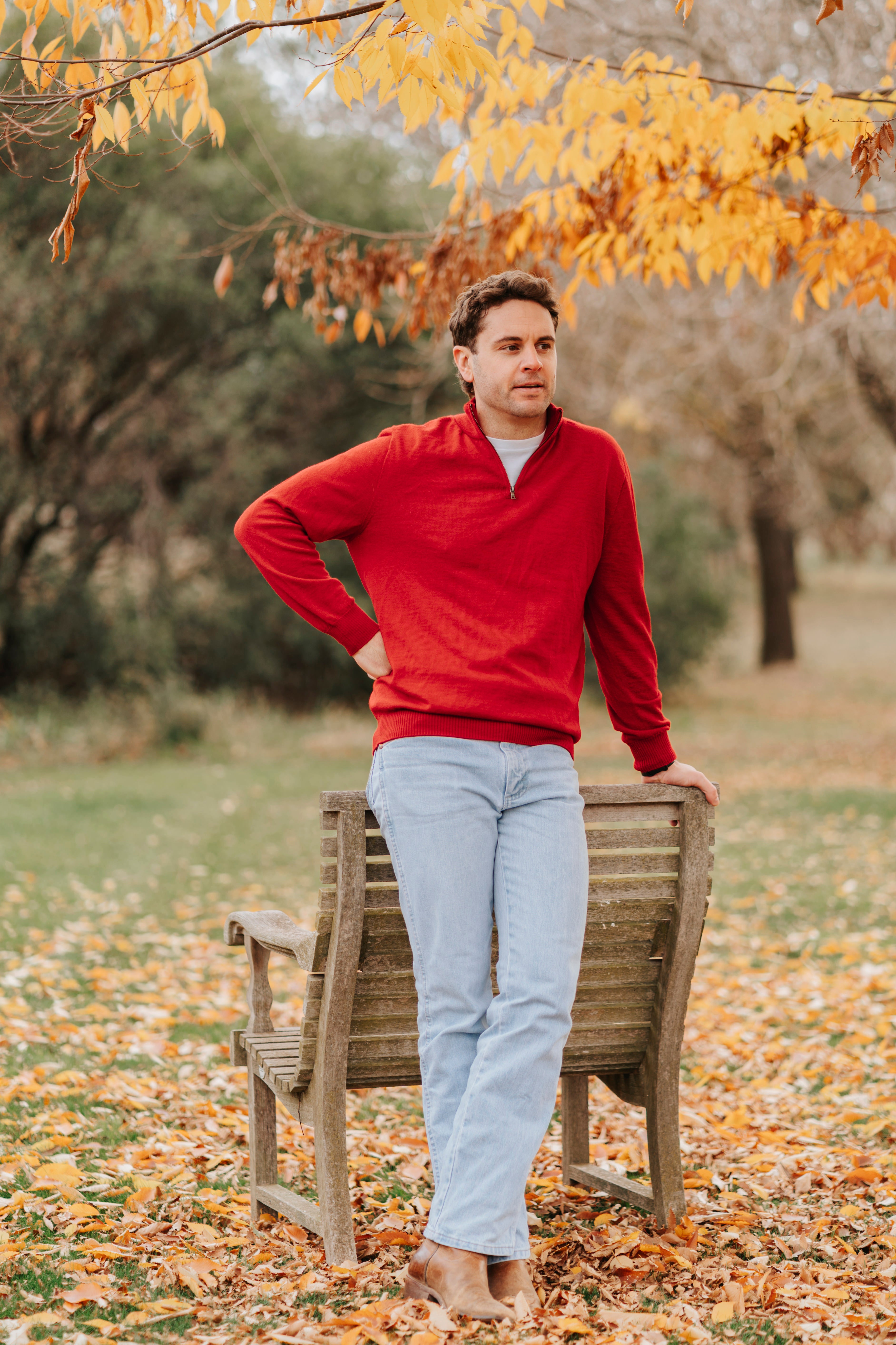 Man in a red merino sweater and light jeans standing on a wooden bench with autumn leaves and trees in the background.