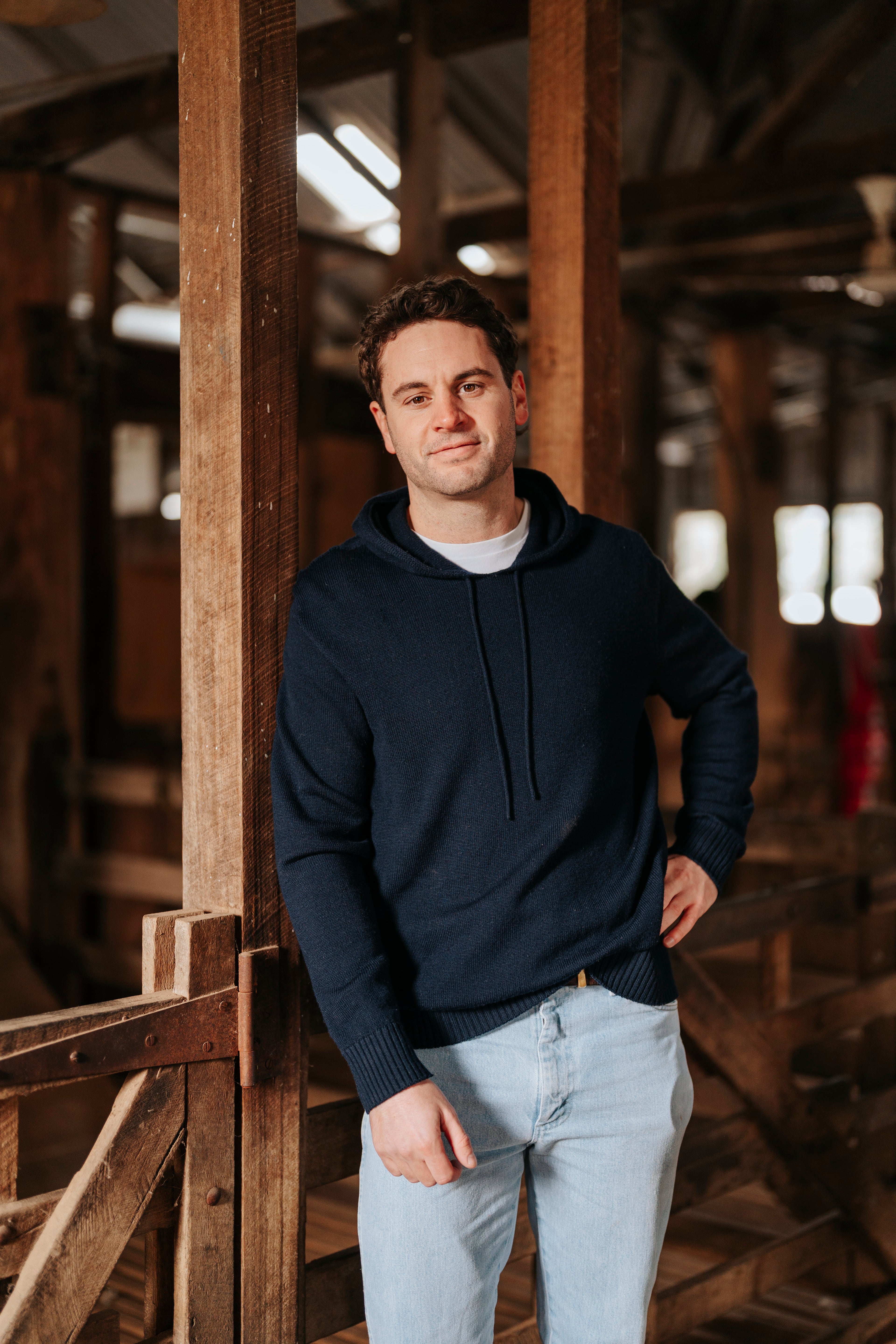 Man wearing a navy hoodie and light jeans standing in a rustic wooden interior.