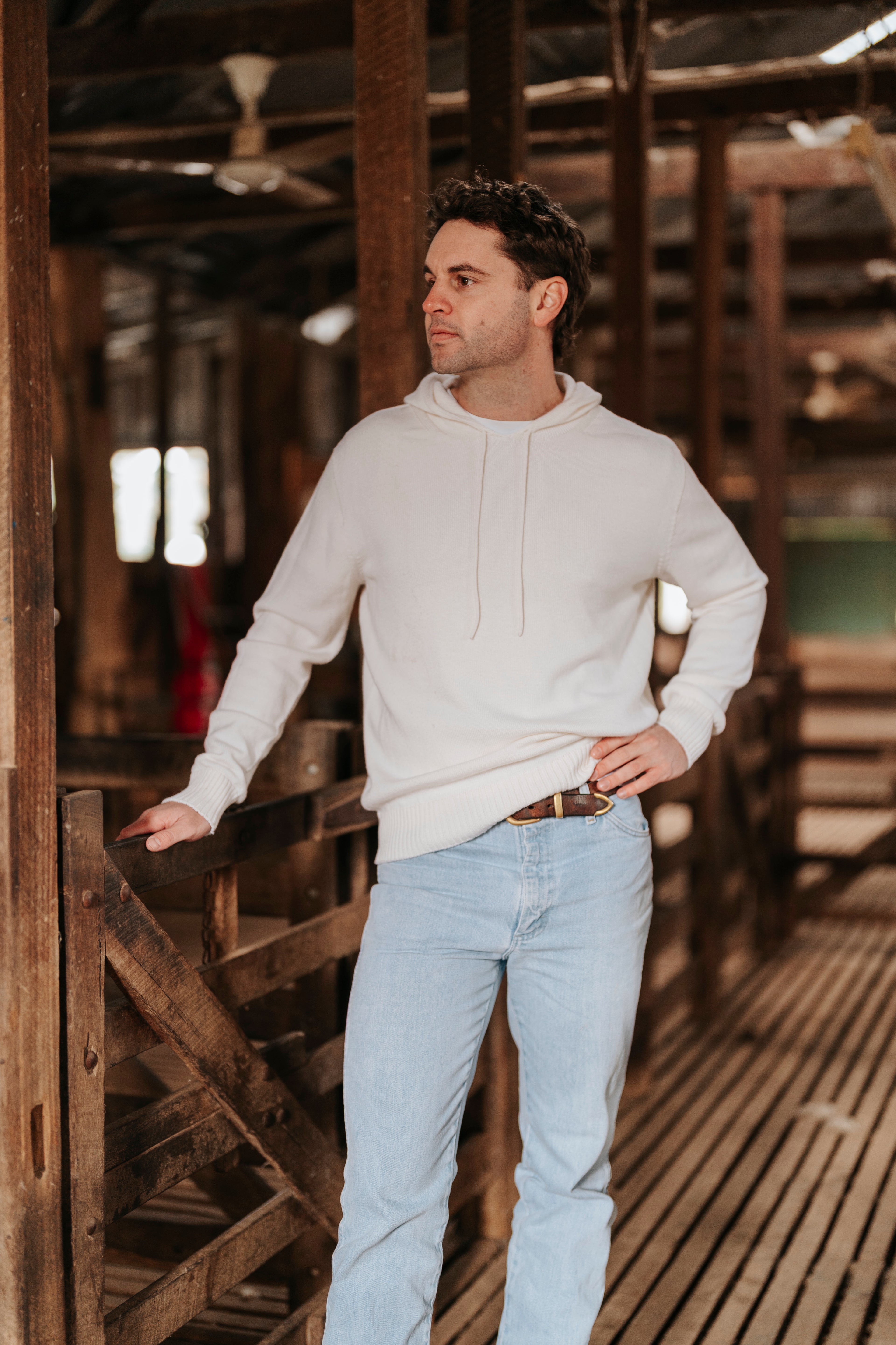 Man wearing a white Merino wool hoodie and light blue jeans standing in a rustic wooden interior.