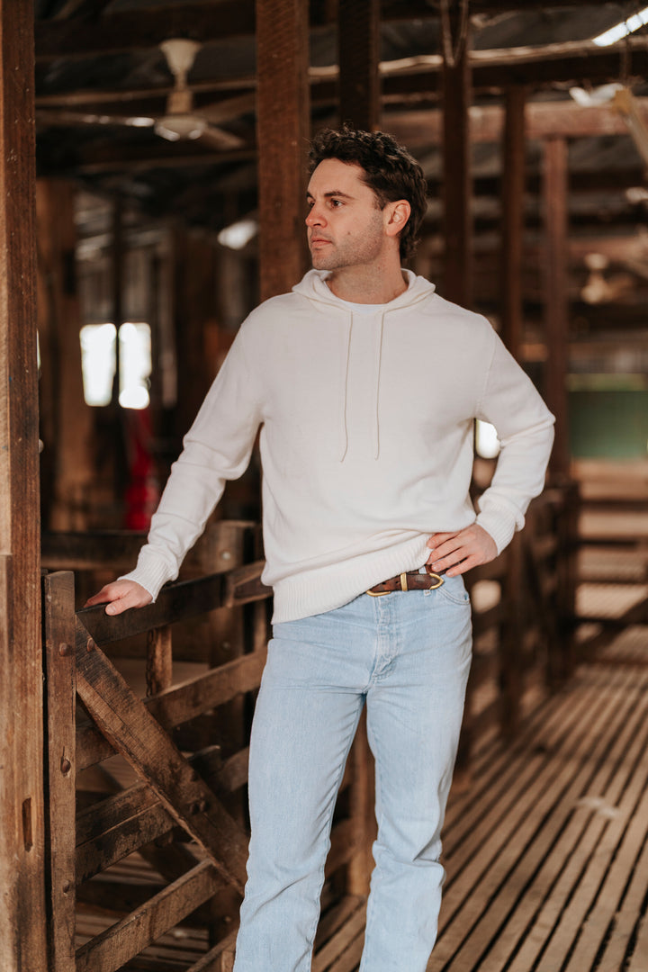 Man wearing a white Merino wool hoodie and light blue jeans standing in a rustic wooden interior.