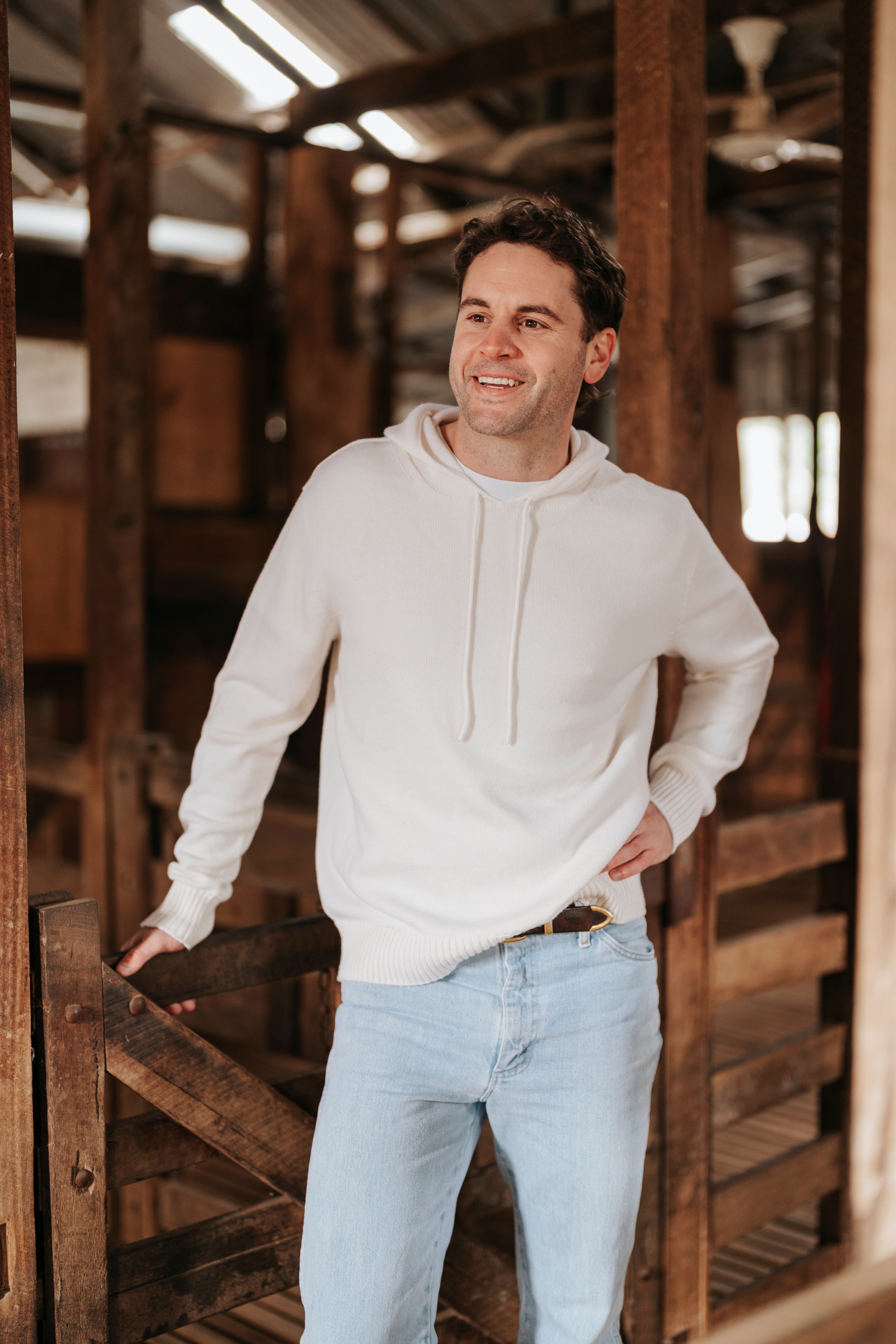 Man wearing a white Merino wool hoodie and light blue jeans standing in a rustic wooden interior.