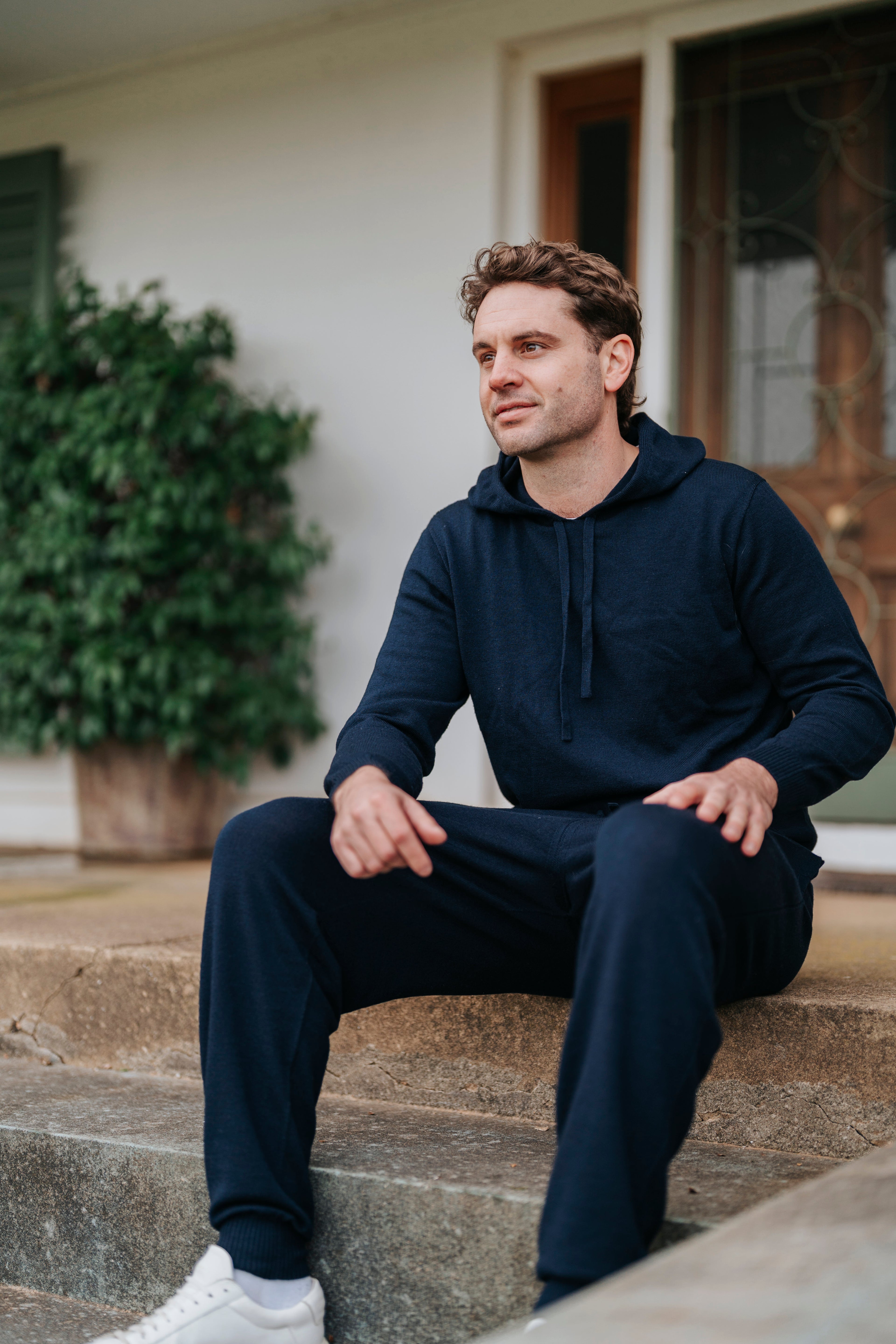 Man sitting on steps wearing a navy blue hoodie and pants with white sneakers.