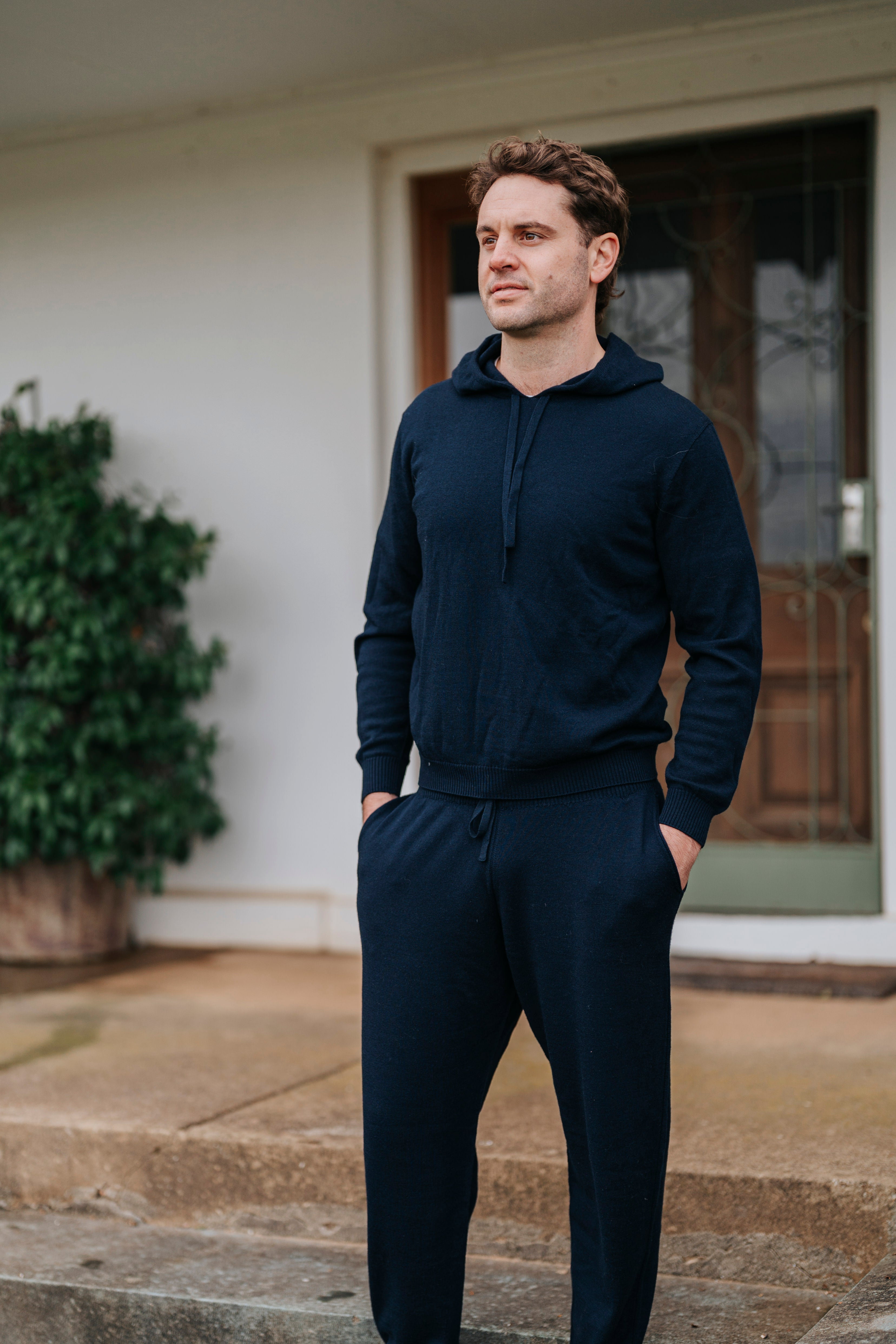 Man wearing a navy blue hoodie and pants standing on a porch.