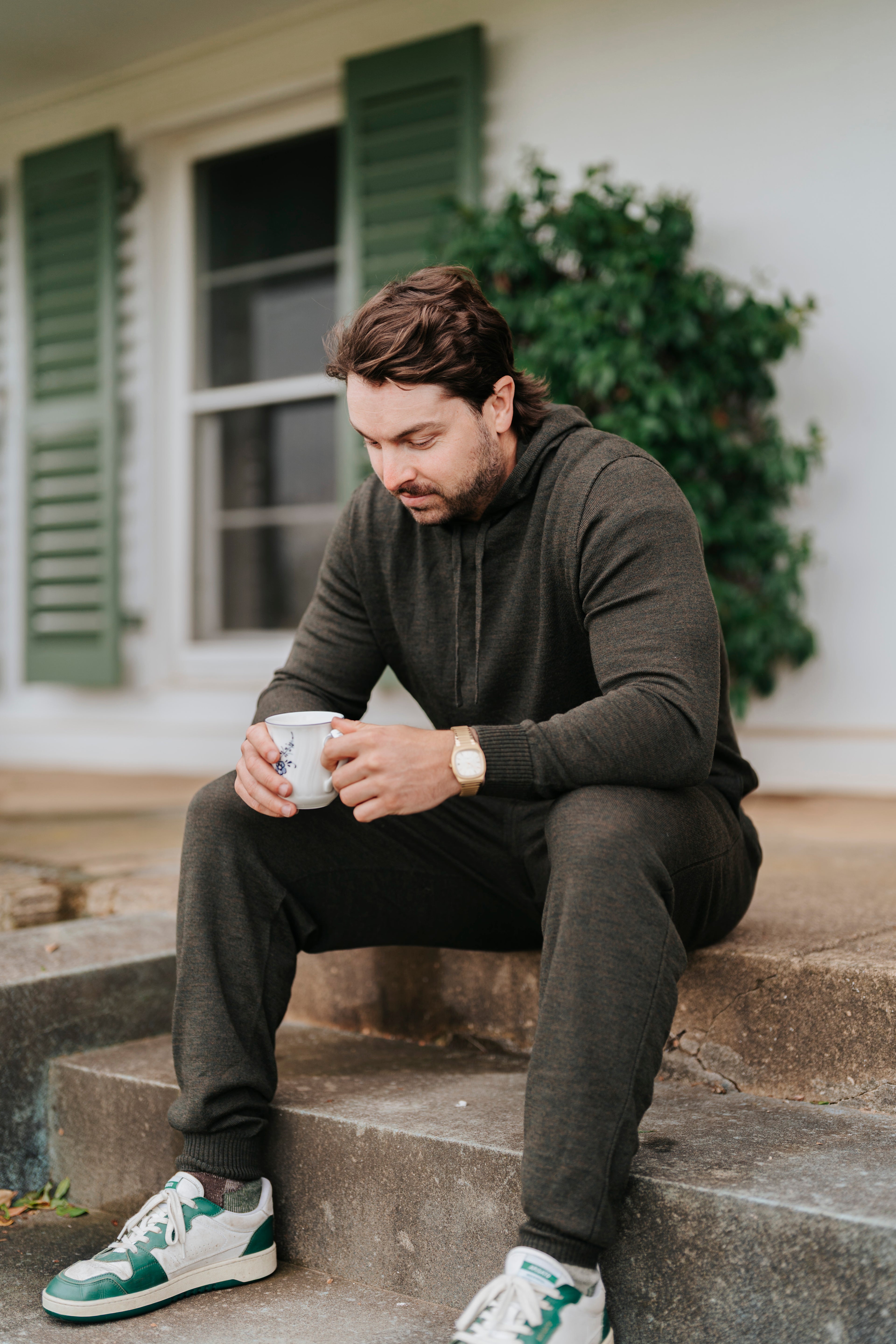 Man sitting on steps holding a mug, wearing a merino sweater and pants with green sneakers.