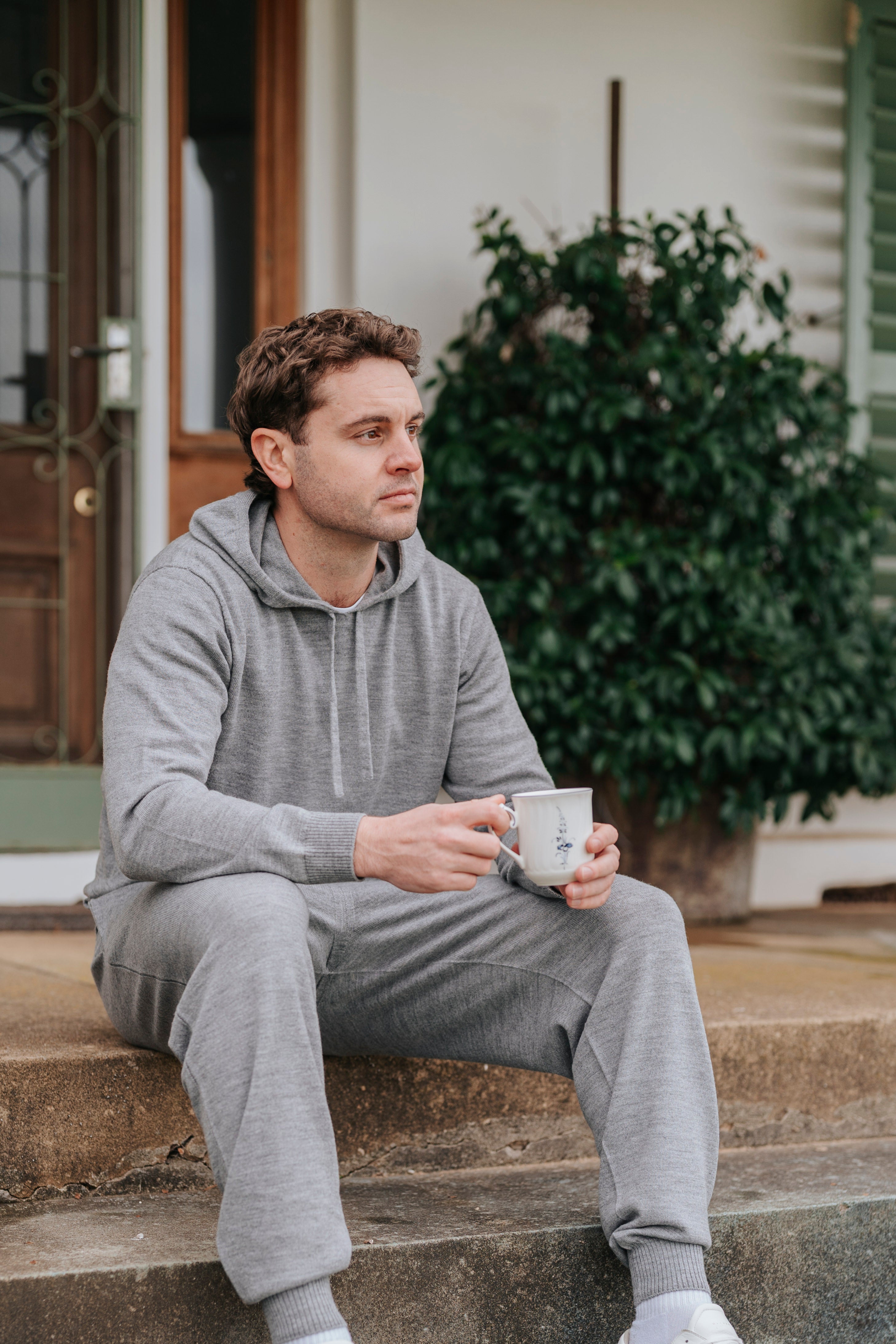 Man in Grey Merino wool Sweatpants sitting on steps holding a mug, with a plant in the background.
