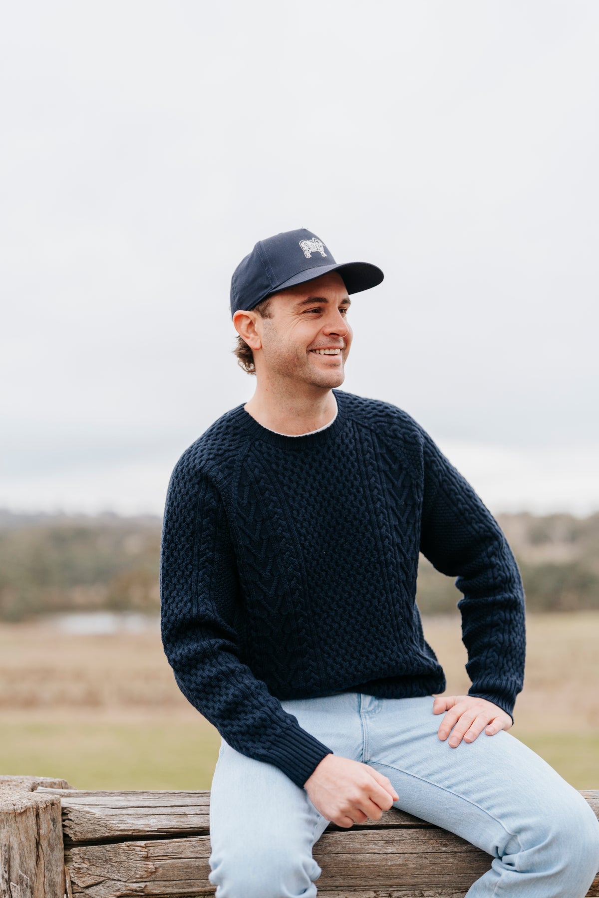 Man wearing a navy sweater and cap sitting on a wooden log with a field and sky in the background #Color_navy