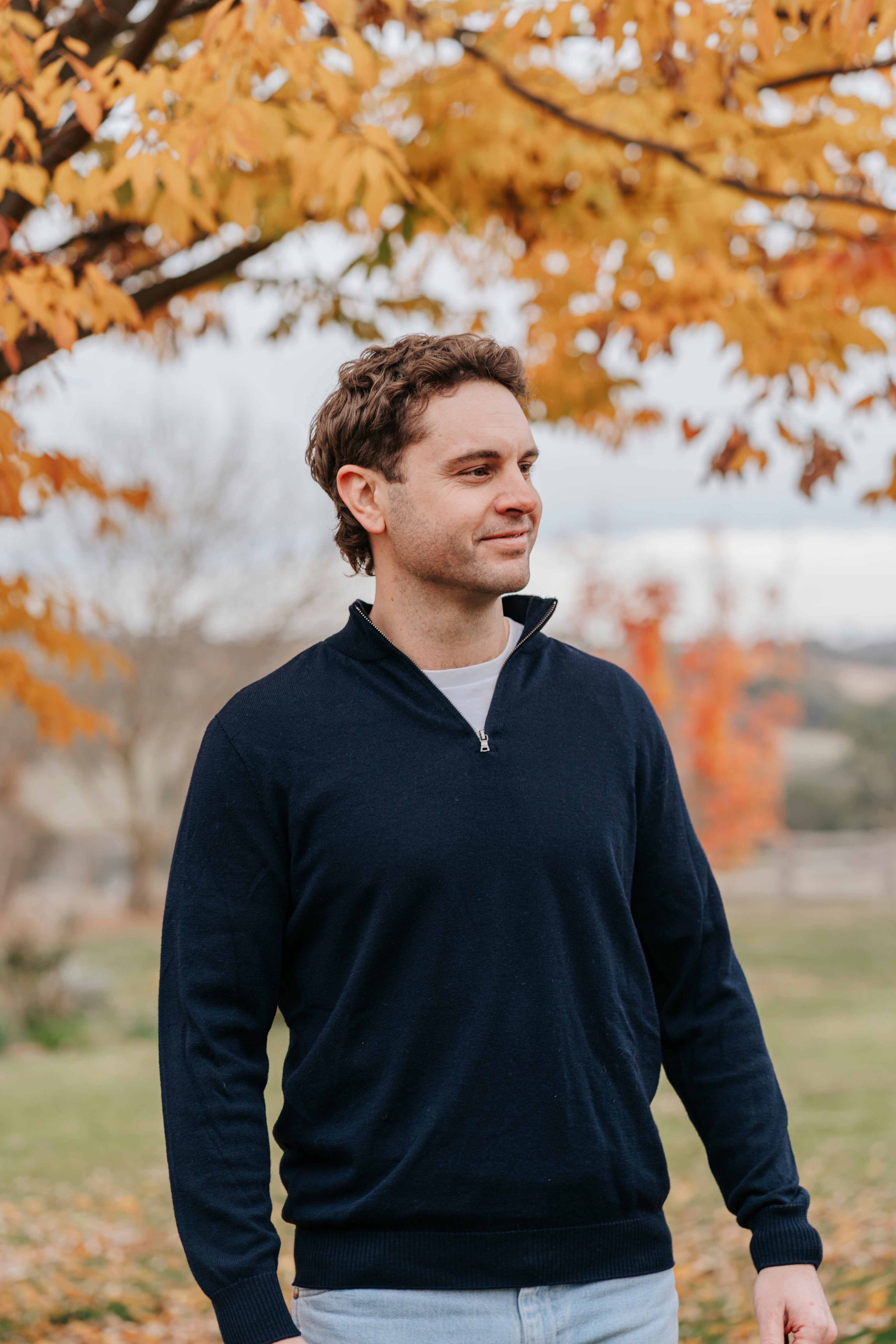Man wearing a navy blue sweater standing in a park with autumn trees in the background