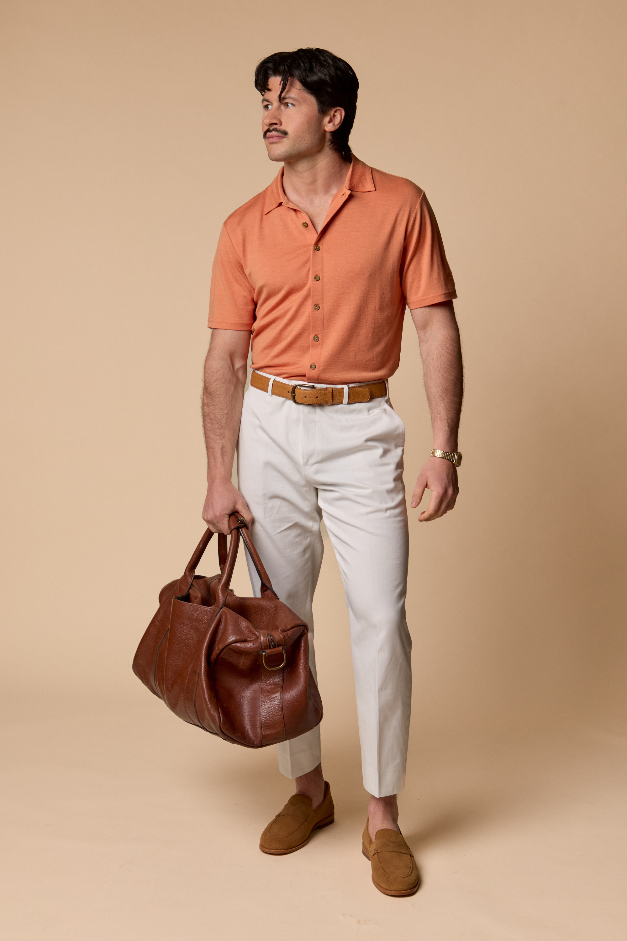Man wearing a Sir W. Merino Bruce Caramel shirt, white pants, and brown shoes holding a brown leather bag