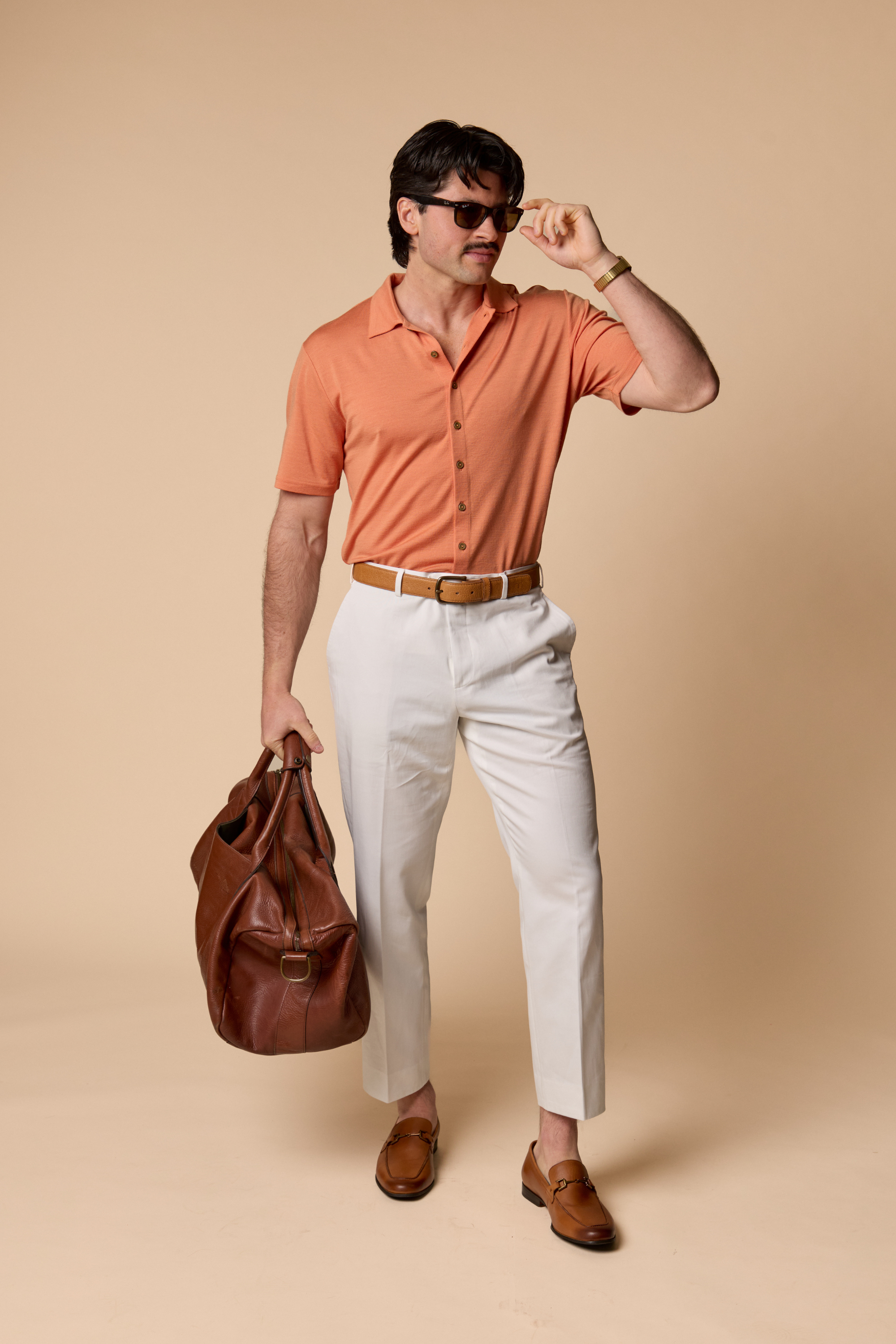 Man wearing a Sir W. Merino Bruce Caramel shirt, white pants, and brown shoes, holding a brown bag against a beige background.