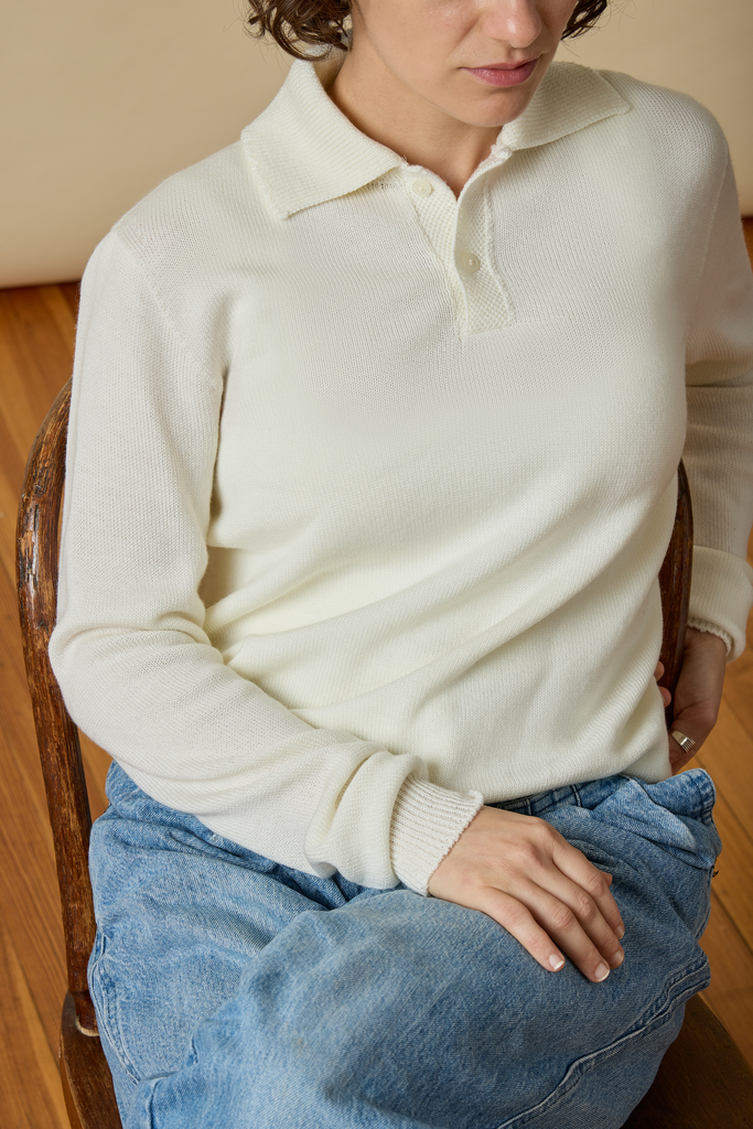 Person wearing a white Sir W. Merino sweater and blue jeans sitting on a wooden floor.