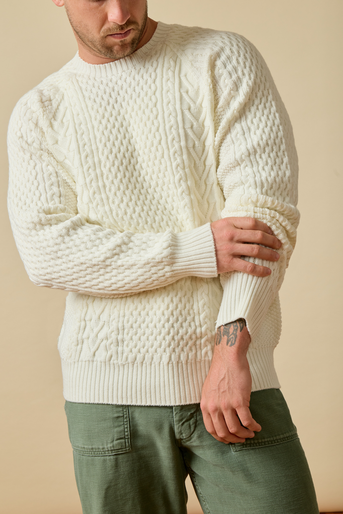 Man wearing a white cable knit Sir W. Merino sweater and green pants against a beige background