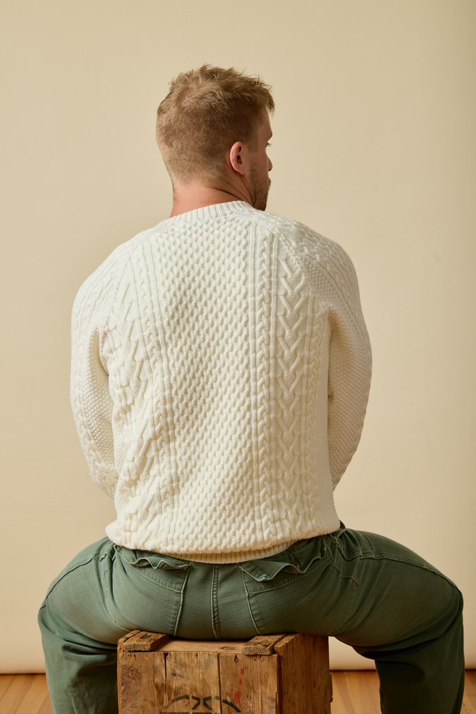 Man wearing a white cable knit Sir W. Merino sweater sitting on a wooden box against a beige wall.