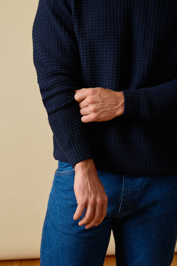 Person wearing a dark blue Sir W. Merino sweater and jeans against a beige wall.