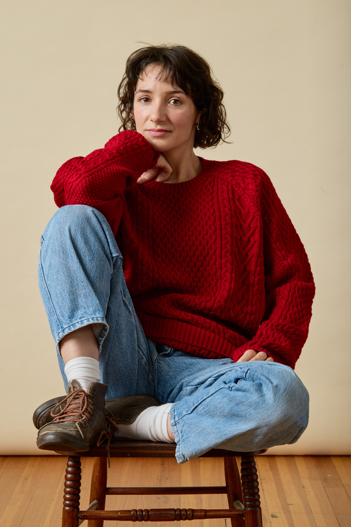 Woman wearing a red Sir W. Merino sweater and blue jeans sitting on a wooden stool against a beige wall.