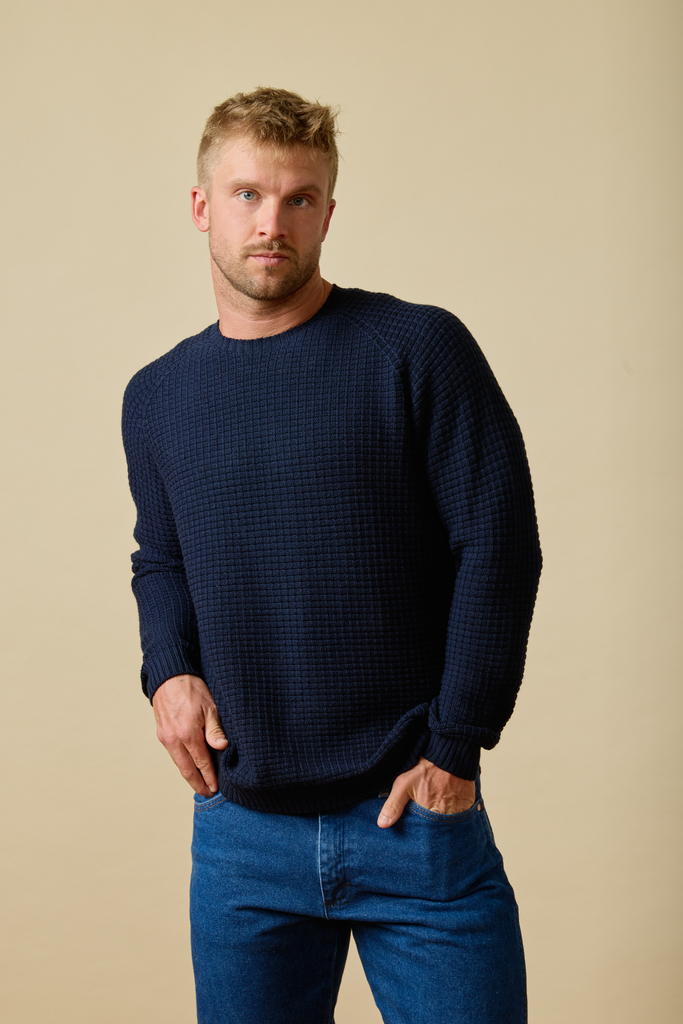 Man wearing a navy blue Sir W. Merino sweater and blue jeans against a beige background