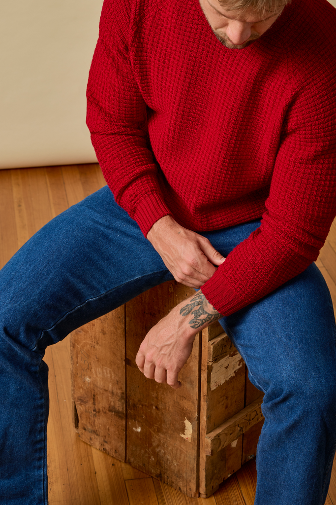 Person wearing a red Sir W. Merino sweater and blue jeans sitting on a wooden box indoors.