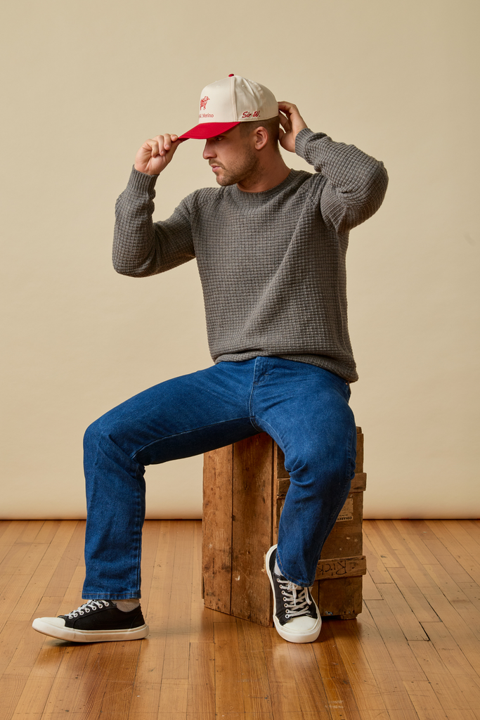 Man sitting on a wooden box wearing a gray Sir W. Merino sweater, blue jeans, and a red and white cap.