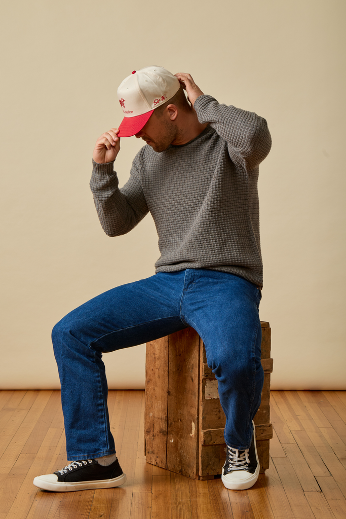 Person sitting on a wooden block wearing a gray Sir W. Merino sweater, blue jeans, and black sneakers with a red cap.