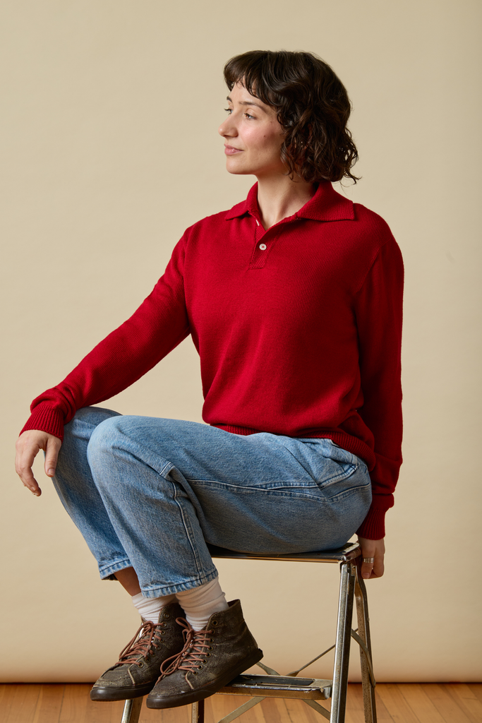 Person wearing a red Sir W. Merino sweater and blue jeans sitting on a stool against a beige wall.