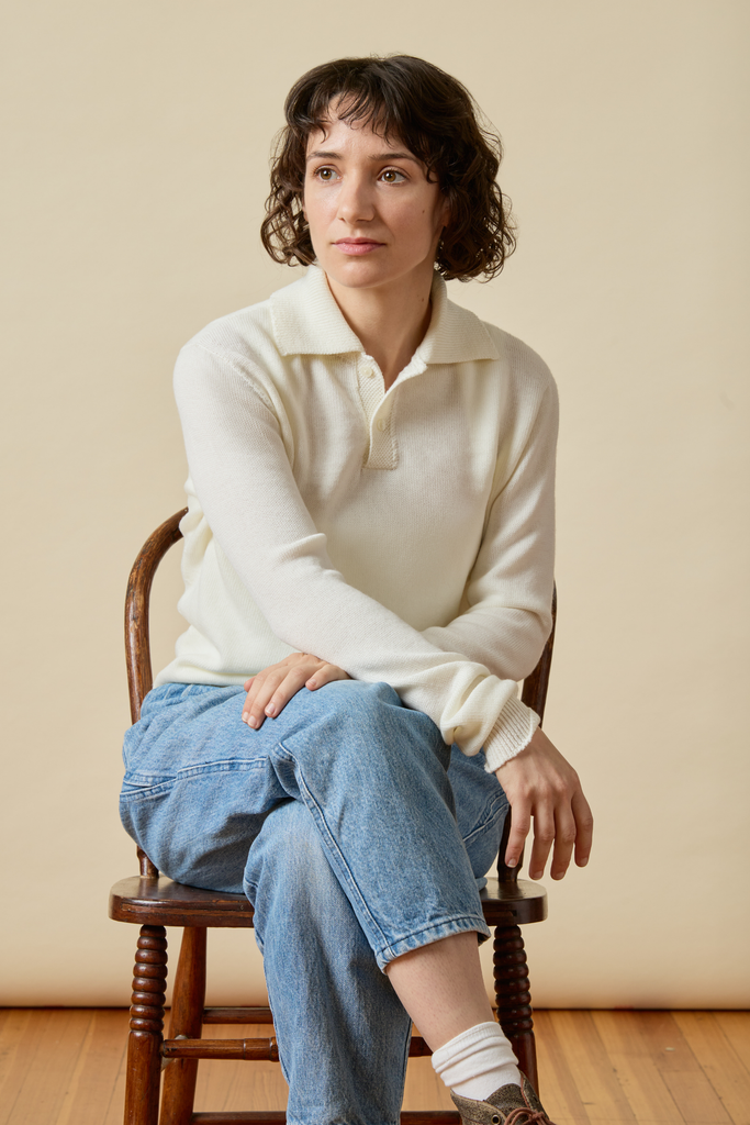 Woman sitting on a wooden chair wearing a white Merino wool sweater and blue jeans against a beige wall.