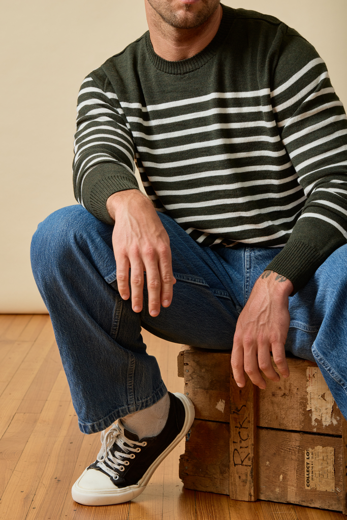 Man sitting on a wooden crate wearing a striped Merino wool sweater and jeans.