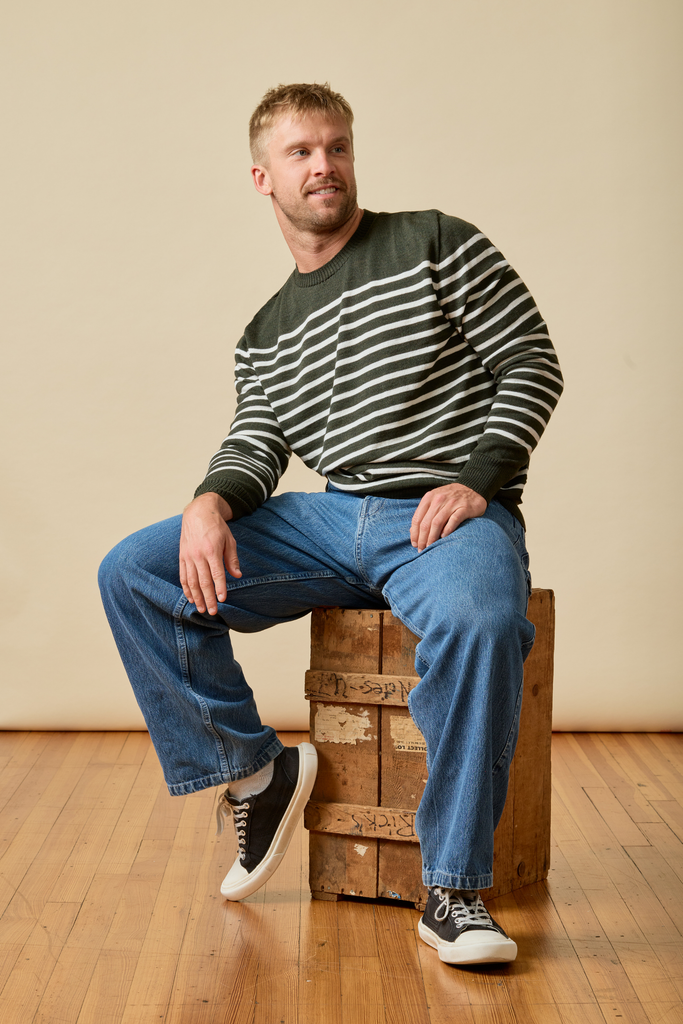 Man sitting on a wooden crate wearing a striped Sir W. Merino wool sweater and jeans.