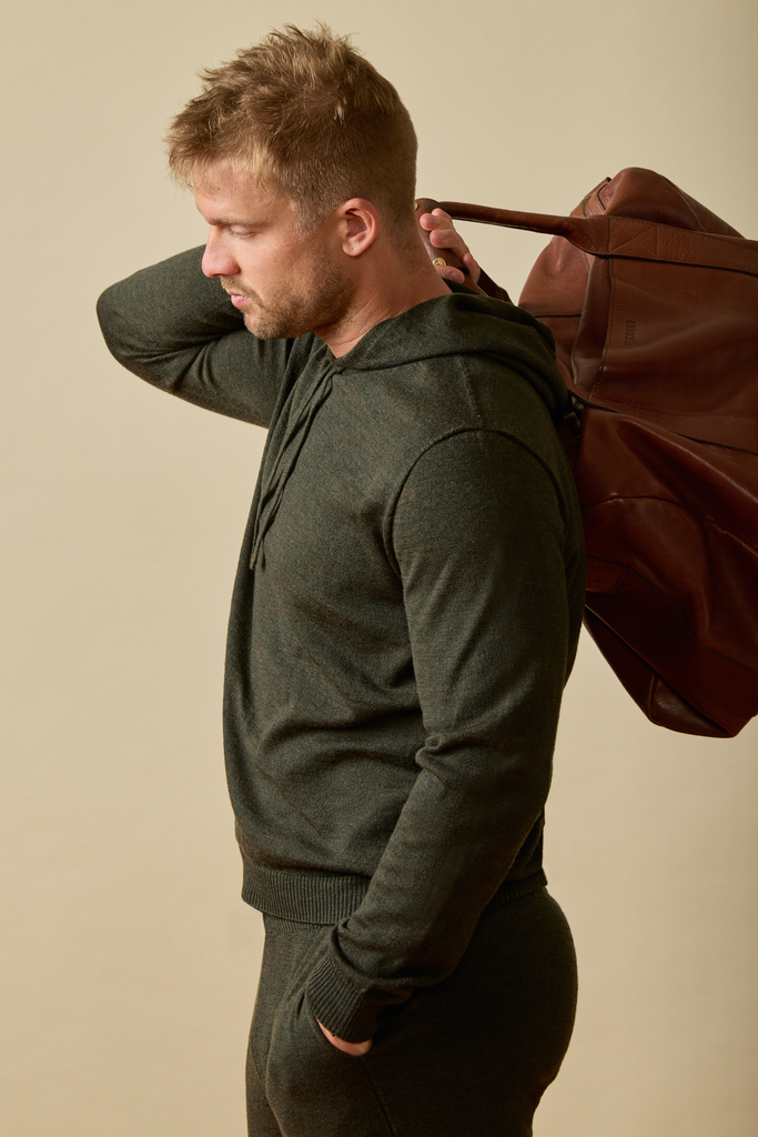 Man wearing a dark green Merino wool hoodie and pants, carrying a brown leather bag on a beige background