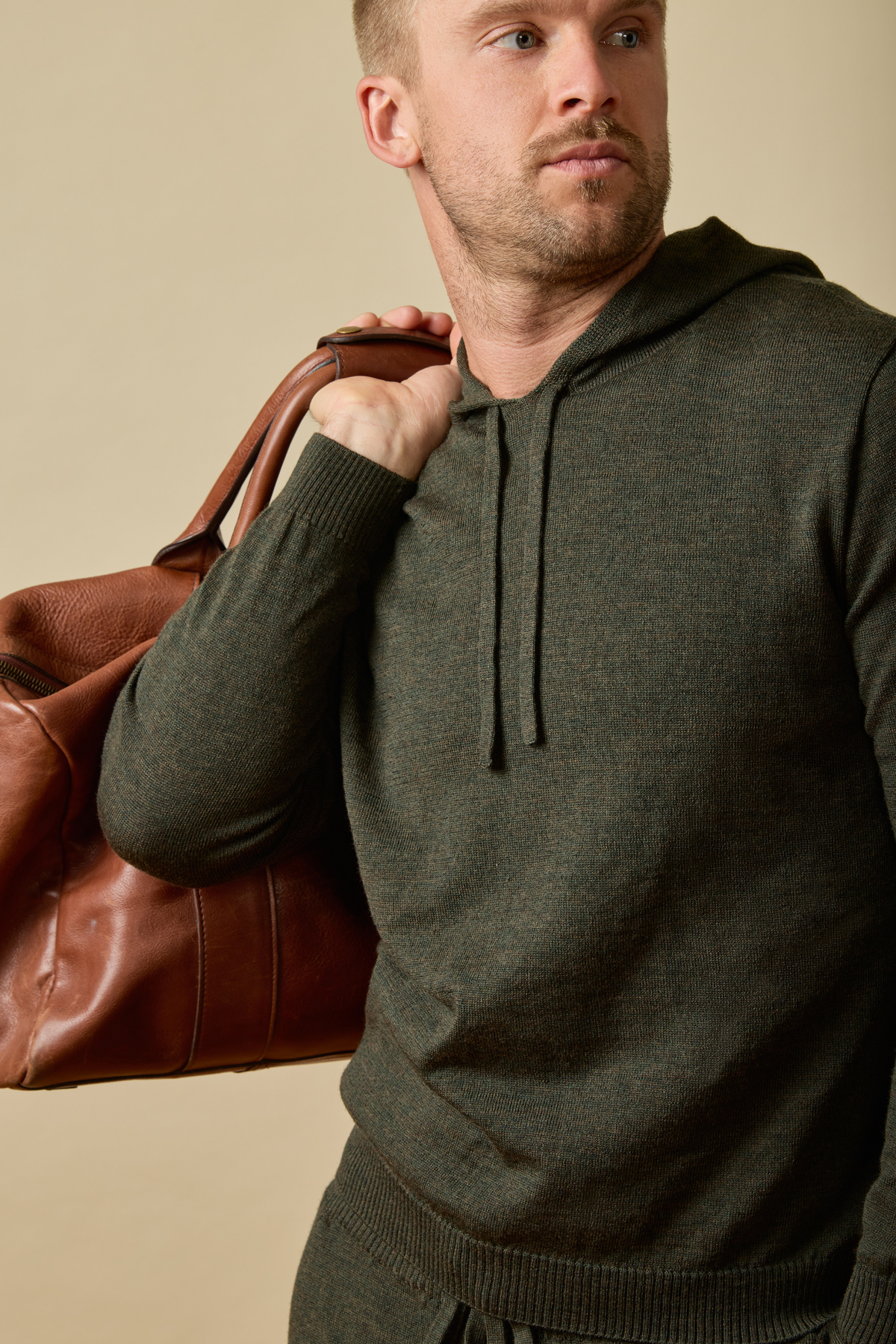 Man wearing a green Merino wool hoodie holding a brown leather bag against a beige background