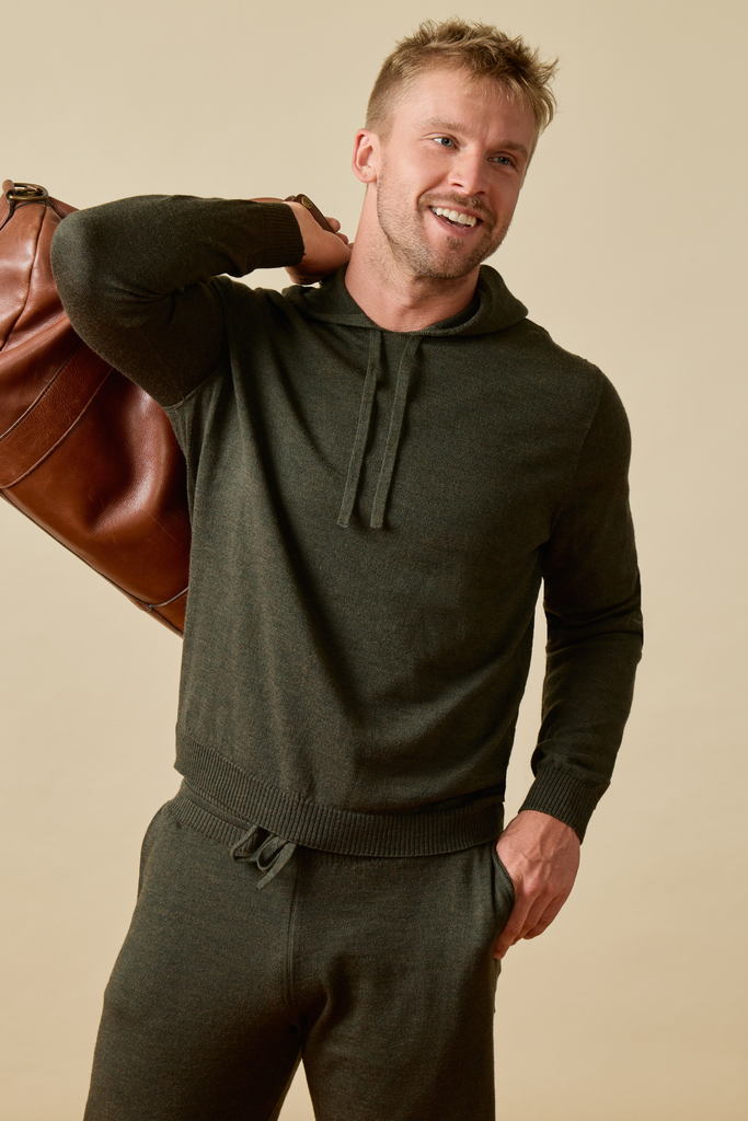 Man wearing a dark green Merino wool hoodie and pants holding a brown leather bag against a beige background