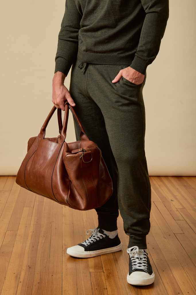 Person holding a brown leather bag wearing a green Merino wool sweater and pants on a wooden floor.