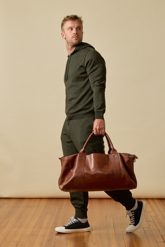 Man holding a brown leather bag in a room with beige walls and wooden floor wearing a dark green Merino wool tracksuit