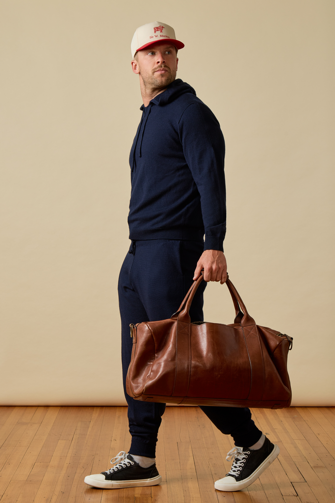 Man wearing a dark navy Merino wool set holding a brown leather bag against a beige wall