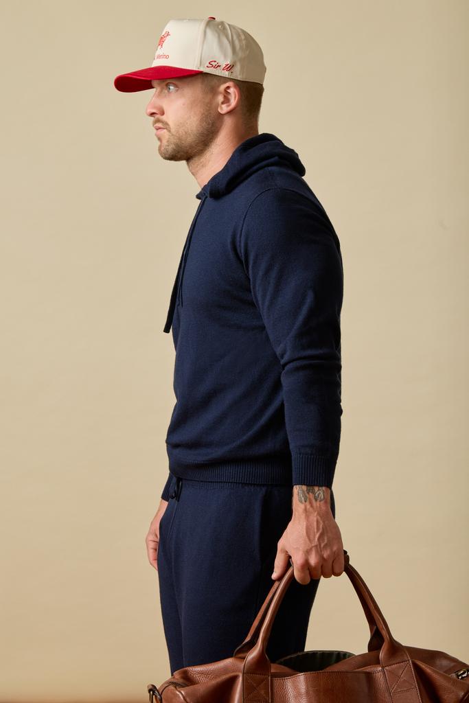 Man wearing a navy Merino wool tracksuit and red and white cap, holding a brown leather bag against a beige background