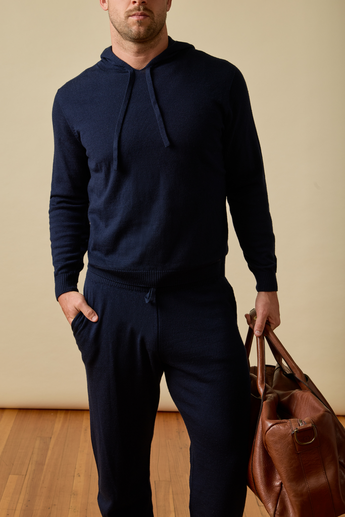 Man wearing a navy blue Merino wool  tracksuit holding a brown leather bag against a beige wall.