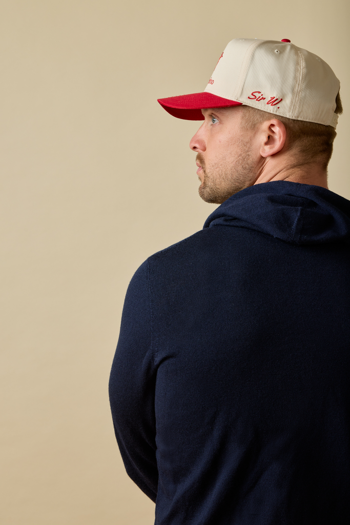 Man wearing a navy Merino wool hoodie and a beige cap with red brim and text, standing against a beige background.