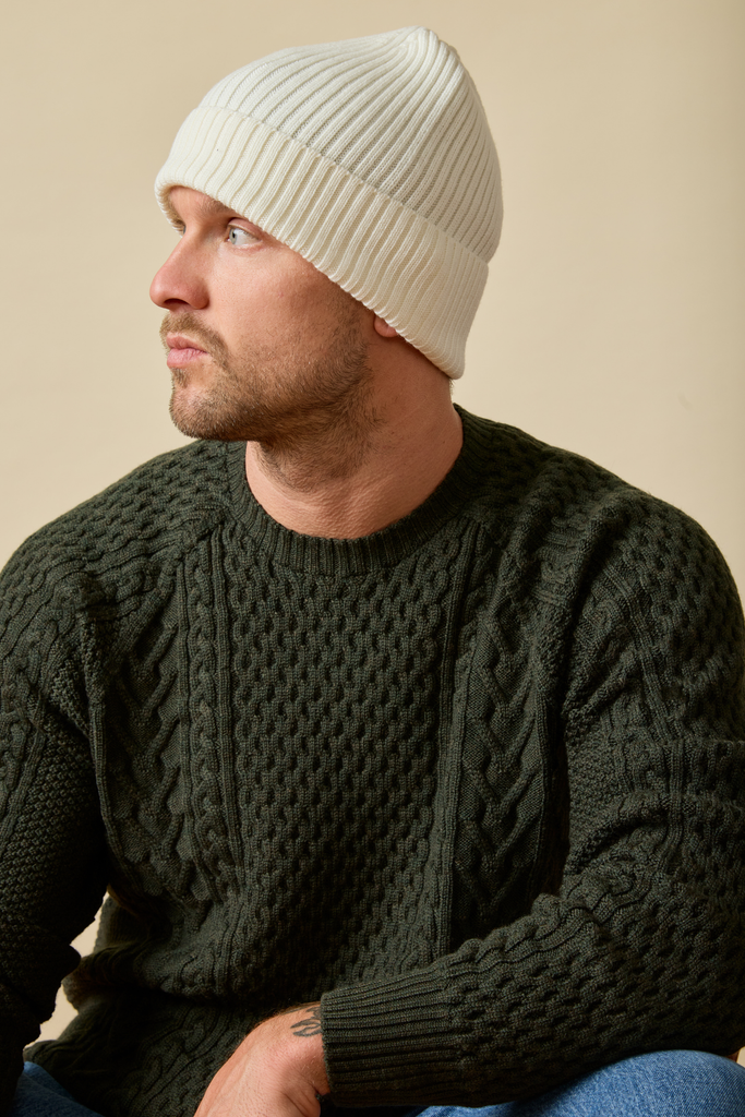 Man wearing a white knit beanie and dark green Merino wool sweater against a beige background