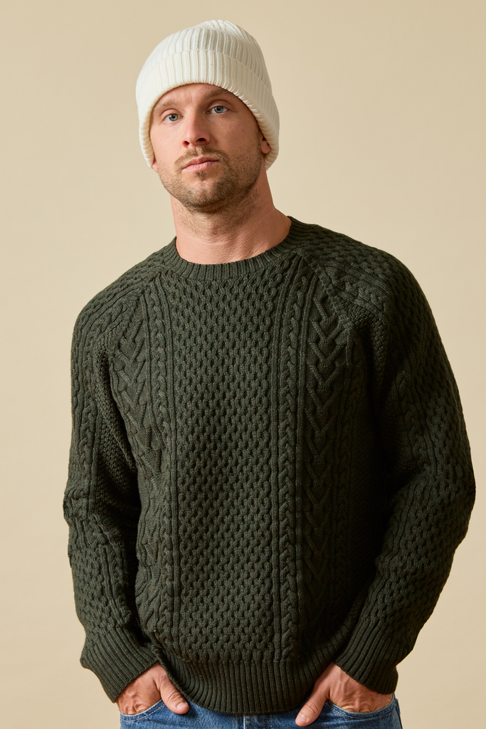Man wearing a dark green Merino wool cable knit sweater and white beanie against a beige background