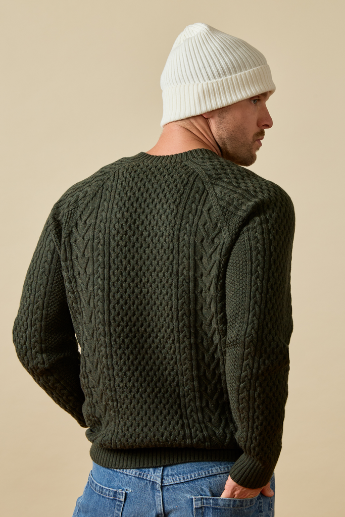 Man wearing a dark green Merino wool cable knit sweater and white beanie against a beige background