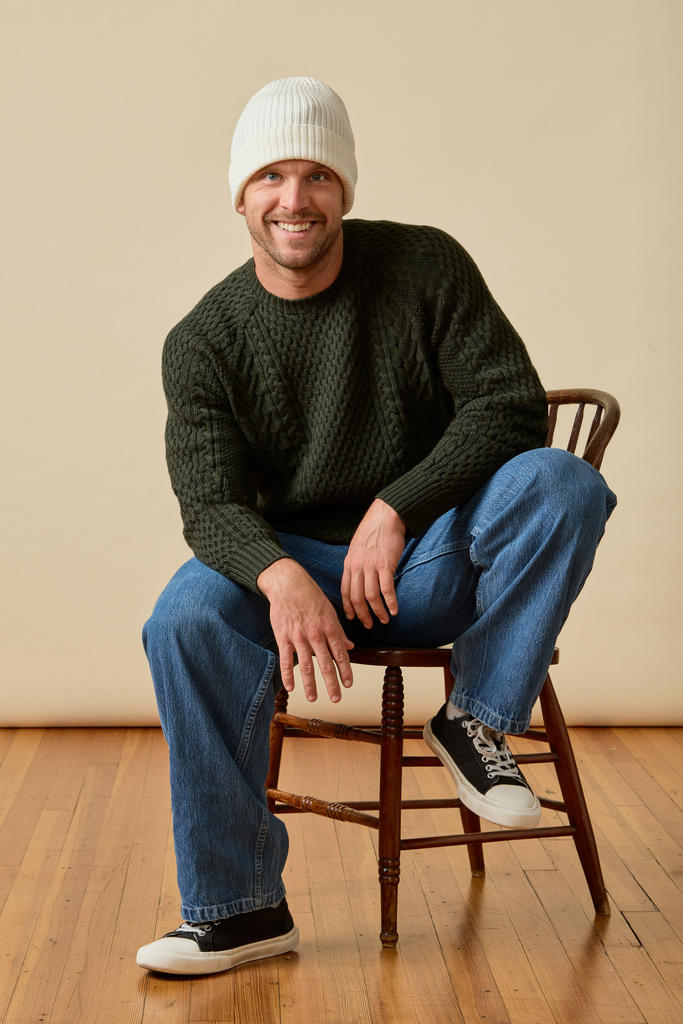 Man wearing a dark green Merino wool cable knit sweater and white beanie against a beige background